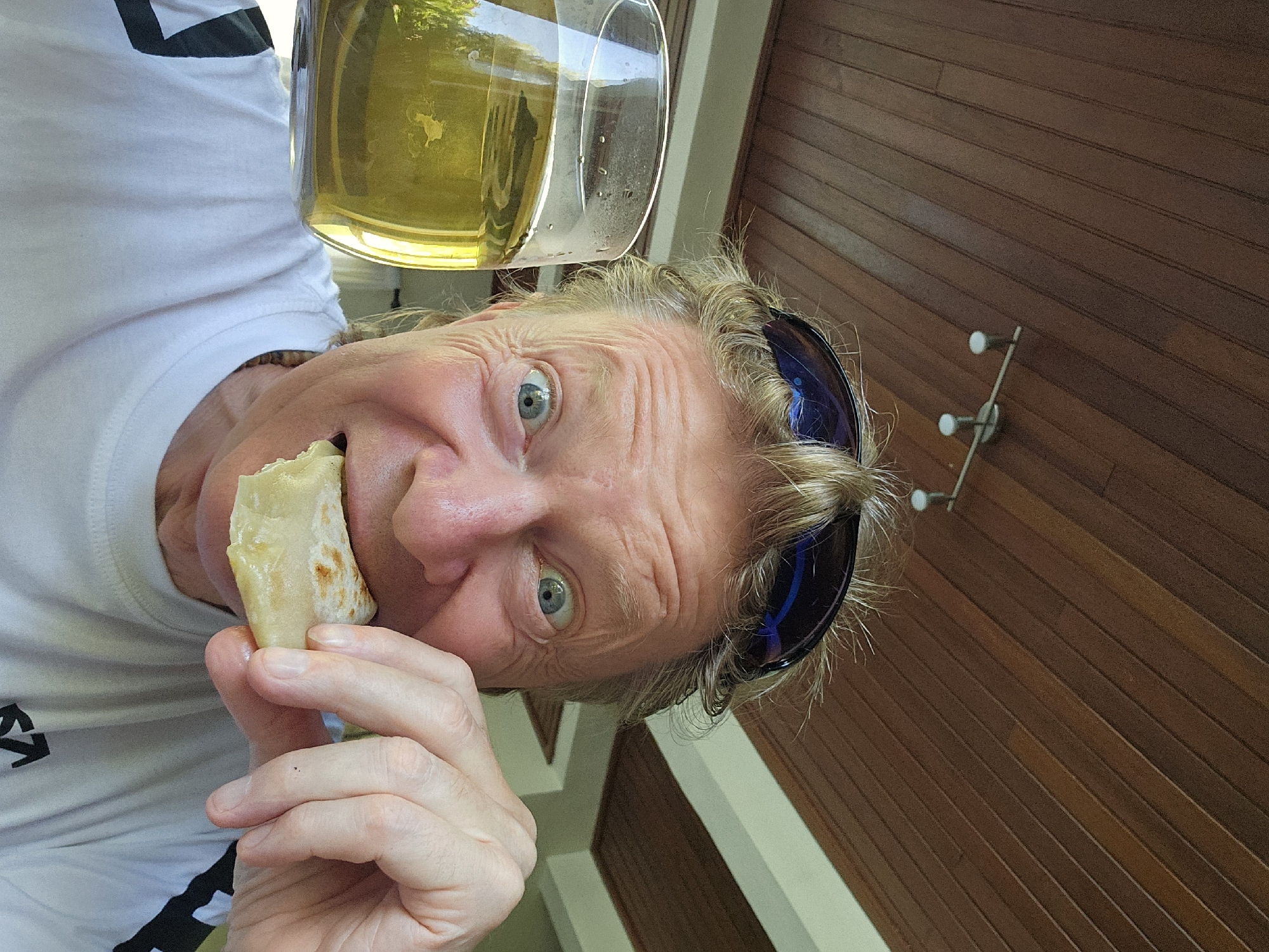 A man is holding a half-eaten burrito in his left hand, near his mouth. He is looking up and to the left at the camera with an open mouth and wide eyes. A glass of beer or cider is over his head. Sunglasses are perched on his head. He is wearing a white t-shirt with black design on the left shoulder. Wood paneling is behind the man, and on the wood paneling is a metal bar with two white knobs. The image may be from Denpasar, Indonesia.