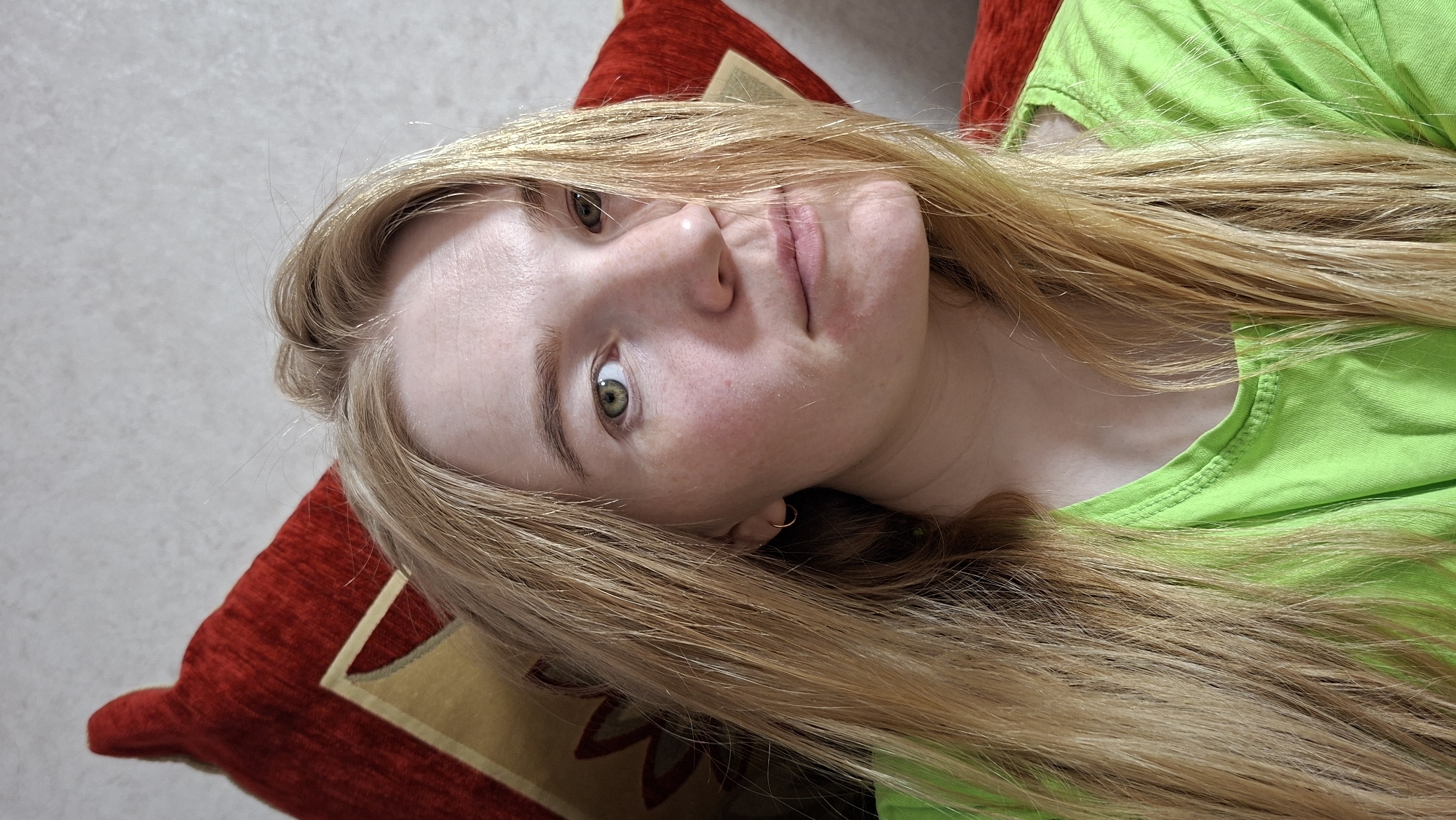 A close-up portrait captures a young woman with long, straight, light blonde hair and fair skin, visibly sprinkled with freckles across her nose and cheeks. She has striking green or hazel eyes that look directly at the camera, and her expression is neutral and relaxed, with a slight hint of a smile.
She is depicted lying down, with her head resting on a plush red cushion that features a gold and beige patterned design. She wears a bright, neon lime green top or t-shirt. A small, simple gold hoop earring is visible in her right ear. Her long hair is spread around her head and over her shoulder, covering parts of the green top. The lighting is soft and even, suggesting an indoor setting. The background behind the cushion appears to be a plain, light-colored wall. There is no visible text within the image.