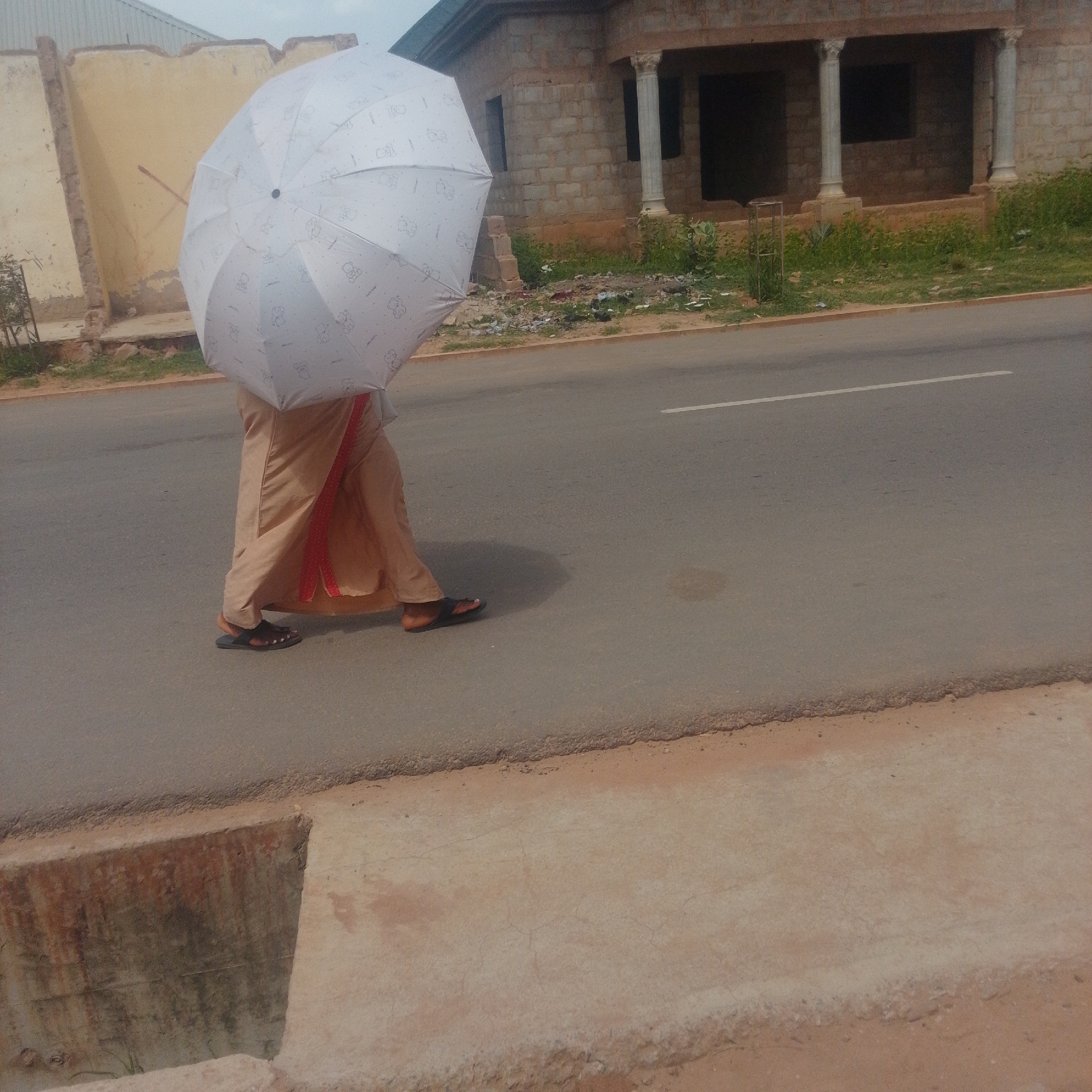 A person walks on a paved road under a white umbrella patterned with cartoon bears. The person is wearing a long, tan garment and dark sandals. Behind them, a building under construction with brick walls and columns is visible, along with overgrown vegetation and scattered debris. The scene appears to be in an urban setting, possibly Gombe, Nigeria, given the architectural style and landscaping. The ground in the foreground is a concrete walkway with visible signs of wear and staining. The road has a white line marking its center.