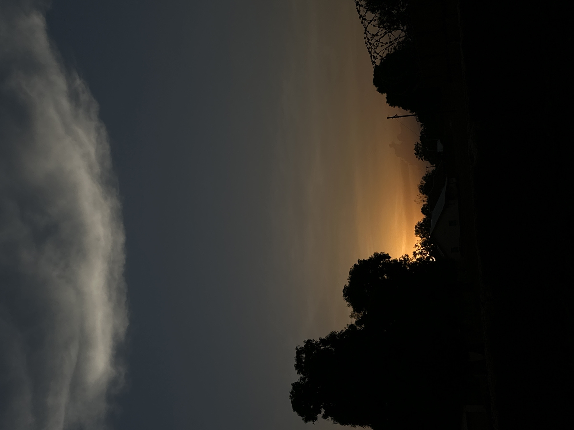 The image depicts a vertical view of an outdoor scene at dusk or dawn, possibly in Bukuru, Nigeria, though this cannot be verified. To the left is a large, vertical column of gray clouds. To the right, the sky transitions from gray to orange, suggesting a sunrise or sunset. Silhouetted against the bright sky are trees and structures, including a building with visible windows. A wire fence is discernible among the silhouetted trees. The right side of the image is in deep shadow, obscuring details. No people are visible.