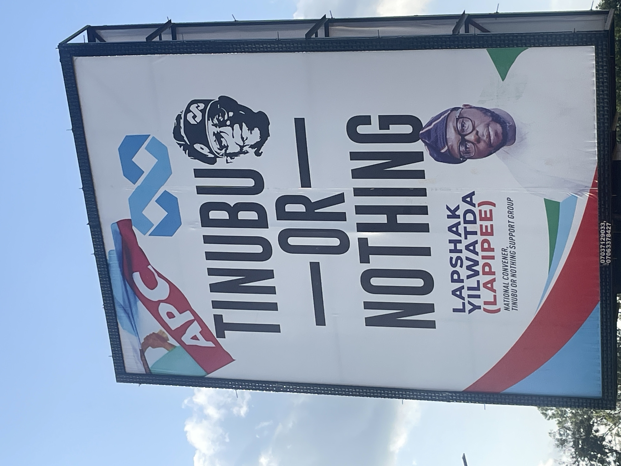 A political billboard is displayed outdoors against a bright blue sky with scattered clouds. The billboard features large black text that reads "TINUBU - OR - NOTHING." Above this text, the letters "APC" are visible on a red and blue striped banner, and an infinity symbol logo in blue is also present. A black and white portrait of a man wearing a cap and glasses is positioned to the right of the "TINUBU" text. Below the main slogan, the name "LAPSHAK YILWATDA (LAPIPIEE)" is written in purple, followed by "NATIONAL CONVENER, TINUBU OR NOTHING SUPPORT GROUP." Below this, a color portrait of a man wearing glasses and a traditional cap is shown. The bottom of the billboard has a strip with the Nigerian flag's colors (green and white) and some text at the very bottom indicating phone numbers. The billboard is framed by a dark metal structure. The image is taken from a slightly low angle, looking up at the billboard. Trees are visible in the background at the bottom left, and a street light pole is also present. The location is identified as Gura Top, Nigeria.