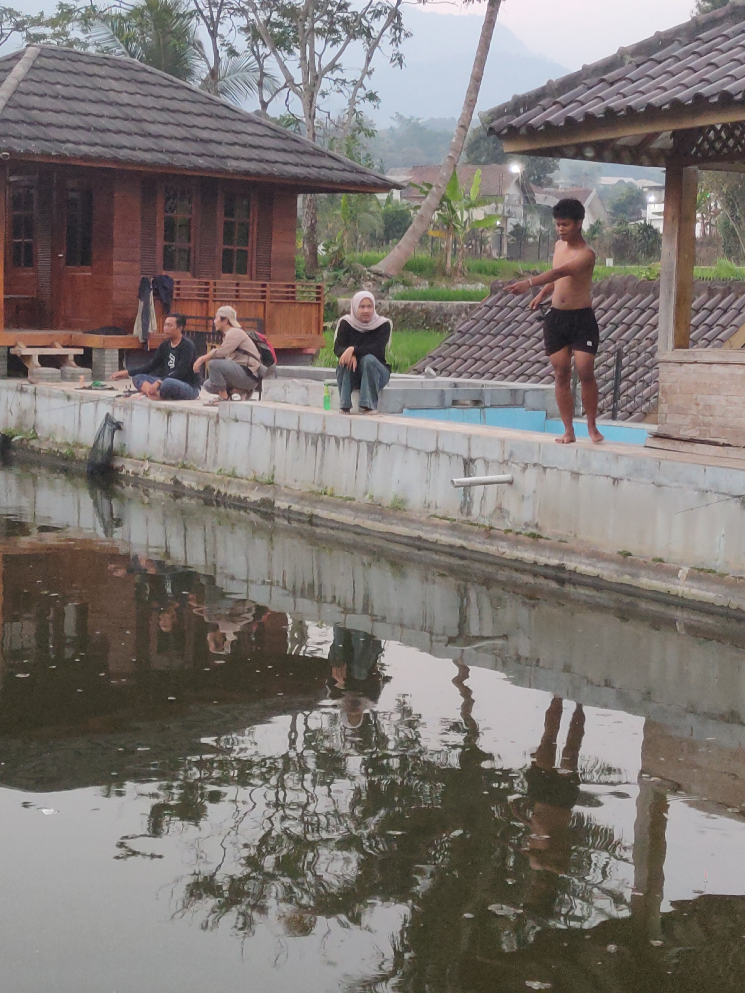 The image depicts a scene outside at dusk, likely in Sumedang, Indonesia. In the foreground, a body of water with reflections of the surroundings dominates the lower half. On the edge of the water, a concrete structure lines the perimeter, where several individuals are present. To the left, three people are seated on the edge of the concrete structure. Two men are seated with their legs crossed, one wearing a dark long-sleeved shirt and the other a light-colored cap and backpack. Between them, a woman in a hijab is also seated. Further to the right, a shirtless young man wearing black shorts stands on the edge, extending his right arm towards the water. Behind the individuals, a wooden building with a tiled roof is visible, along with trees and vegetation. The overall atmosphere suggests a relaxed, outdoor setting.
