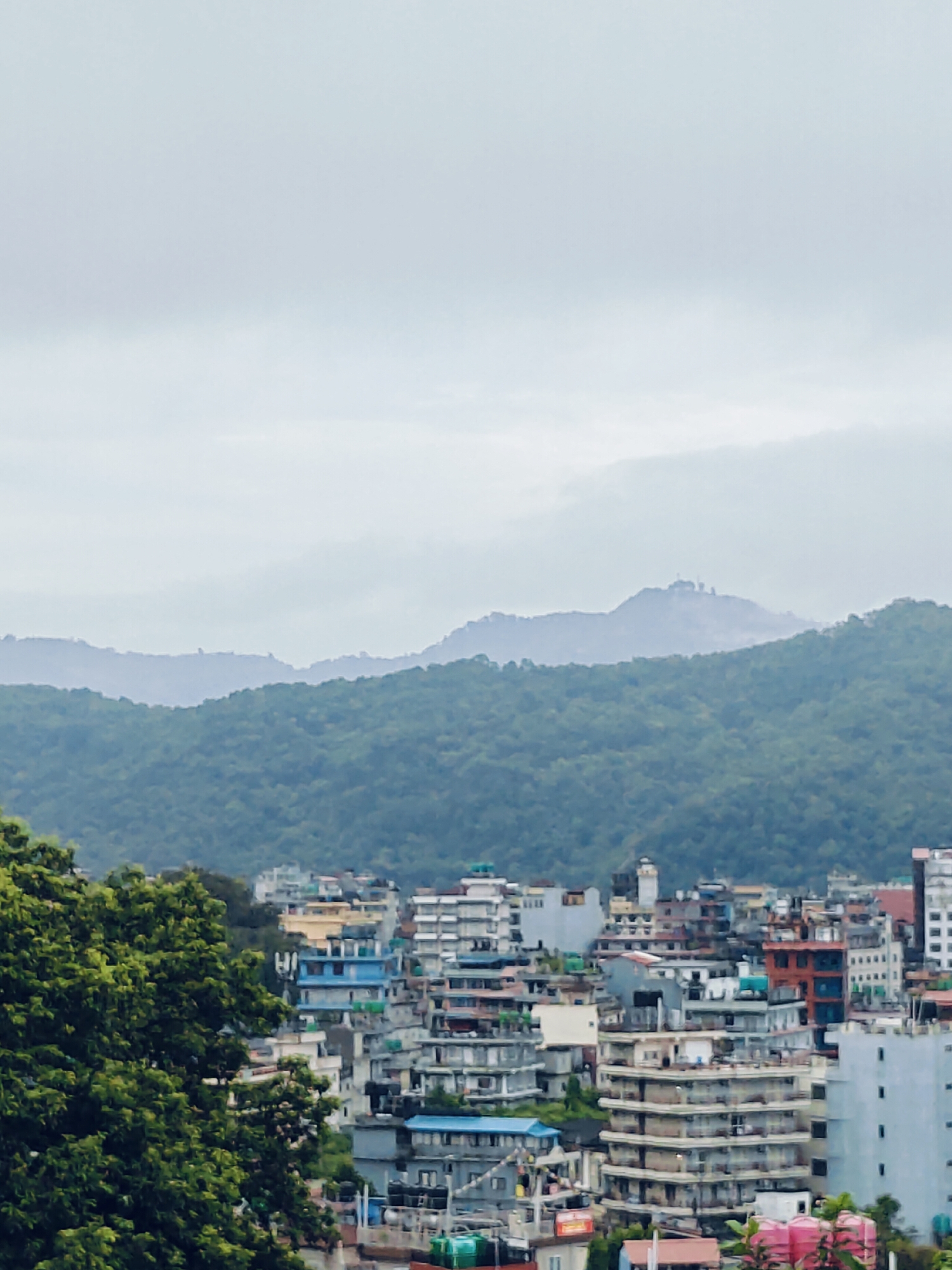 The image captures an elevated view of the city of Pokhara, Nepal, showcasing a dense urban landscape nestled against a backdrop of verdant, rolling hills. In the foreground, lush green foliage, likely from trees, partially obscures the view to the left. The city itself is a mosaic of buildings, varying in height and architectural style, with a predominantly muted color palette of whites, grays, blues, and some warmer tones like yellow and orange. Many of the buildings appear to be multi-story residential or commercial structures.
The middle ground is dominated by the densely packed city, with structures tightly clustered together. Further in the background, the landscape transitions to steep hills covered in dense green forest. The atmosphere appears hazy or overcast, with the distant hills fading into a lighter, muted gray sky. On the furthest ridge, a few structures, possibly antennas or observation points, are faintly visible on the crest of one of the hills. The overall impression is of a busy city integrated into a hilly, natural environment. No people are discernible in the image.