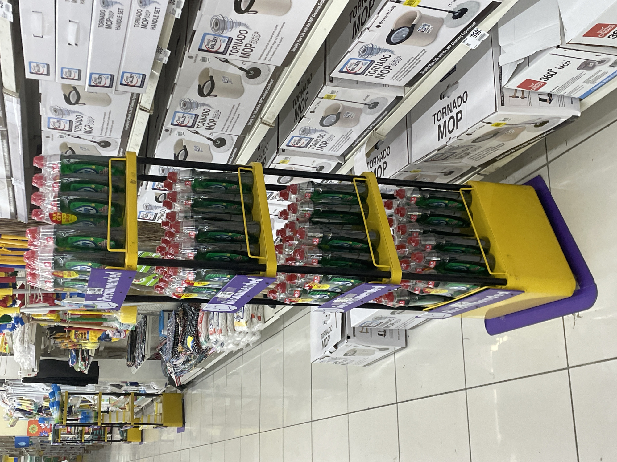 The image shows a retail store aisle, likely a supermarket or department store, in Davao City, Philippines. The aisle features a prominent display of cleaning products, specifically dish soap bottles arranged on yellow tiered shelves. Adjacent to the dish soap display, boxes of "Tornado Mop" cleaning equipment are stacked on higher shelves. The floor is tiled in a white grid pattern. The overall environment suggests a commercial setting stocked with household goods.