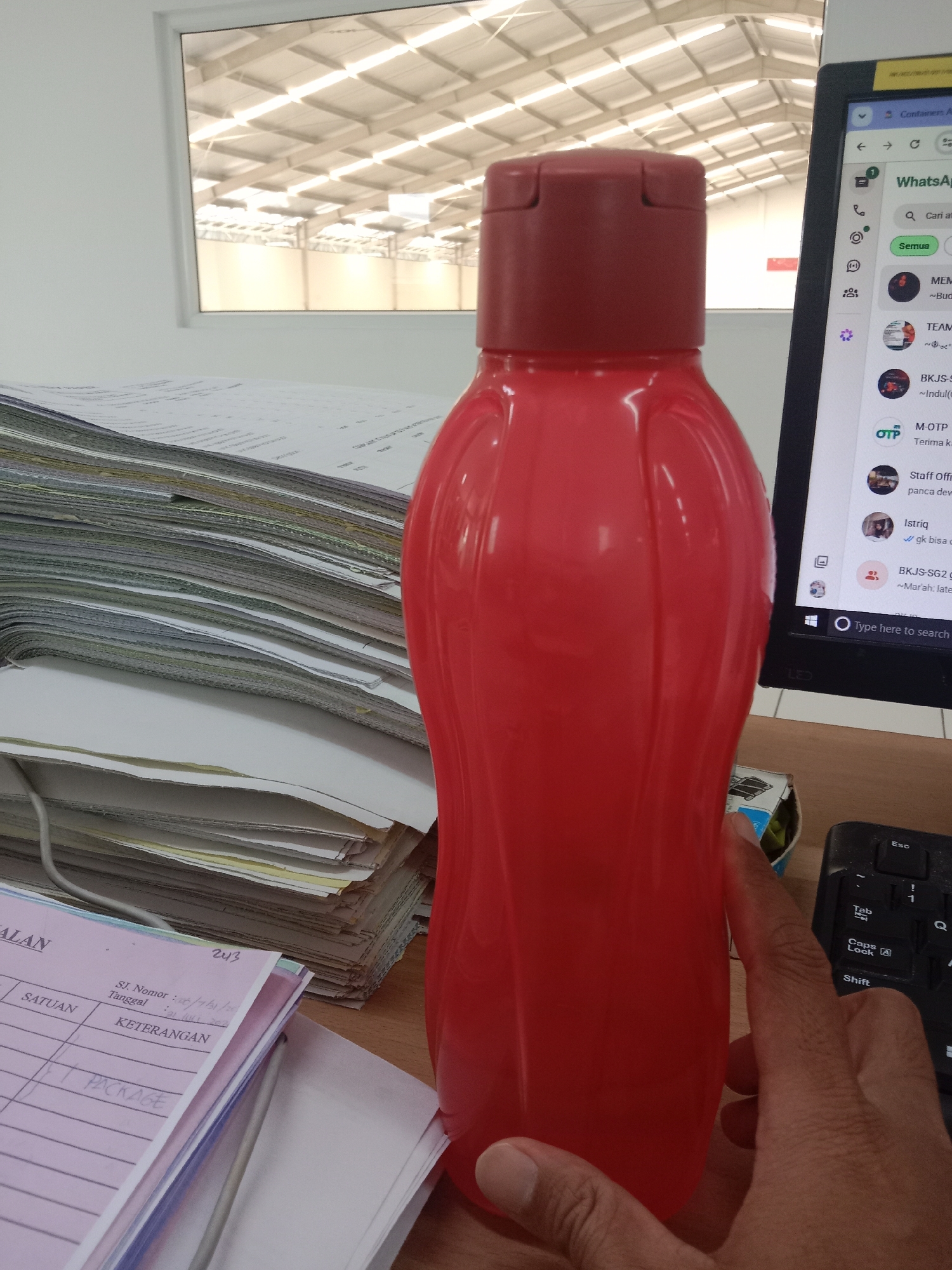 In an indoor setting, likely an office in Semarang, Indonesia, a vibrant red plastic water bottle stands prominently on a wooden desk. The bottle is partially obscured by a stack of papers and a hand reaching towards it. To the right of the bottle, a laptop screen displays a WhatsApp interface, with various chat windows visible, including one labeled "WhatsApp" and a search bar. The background features a large window revealing a brightly lit industrial-style ceiling with rows of lights. The overall impression is one of a workspace, with the water bottle suggesting a moment of personal use within a professional environment.