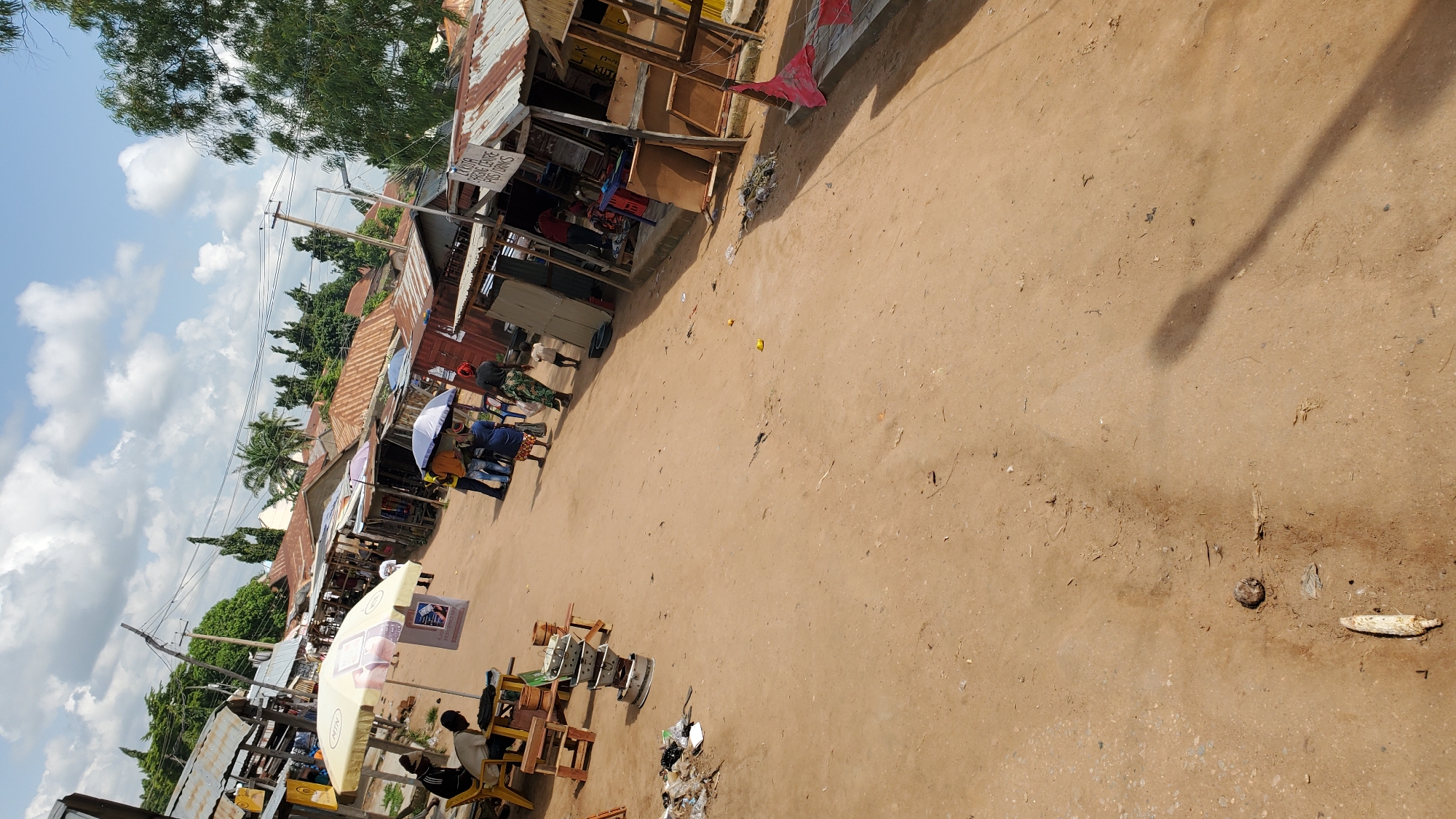 This image depicts a street scene in Jalingo, Nigeria, likely a market area, characterized by a dirt road and numerous market stalls. In the foreground, the sandy ground shows some debris and shadows from overhead objects, including what appears to be a light post.
The middle ground features a collection of open-air market stalls, constructed with corrugated metal roofing and wooden or metal frames. Several individuals are present, some appearing to be vendors and customers. One stall prominently displays an umbrella advertising "MTN". Other stalls have goods displayed, including what might be food items and various products. People are seen standing, walking, and possibly engaging in transactions. One group of people is gathered under an umbrella.
In the background, more market structures and some trees are visible, along with utility poles and power lines, suggesting the presence of basic infrastructure. The sky is partly cloudy, indicating an outdoor daytime setting. The overall impression is a vibrant, albeit simple, marketplace environment in a Nigerian city.