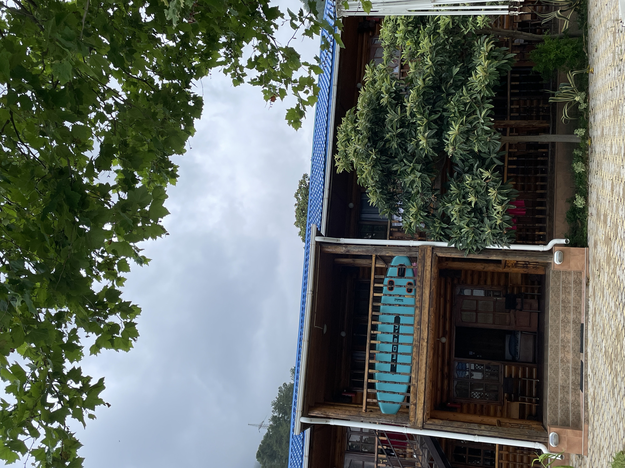 In Abaata, Abkhazia, a wooden log cabin with a blue tiled roof stands under a cloudy sky. A teal-colored paddleboard with black lettering is propped on the railing of the second-story balcony. The cabin has multiple levels with wooden balconies and staircases. Green trees and foliage surround the building, and a cobblestone path leads to the entrance. The scene appears to be a resort or a vacation rental.