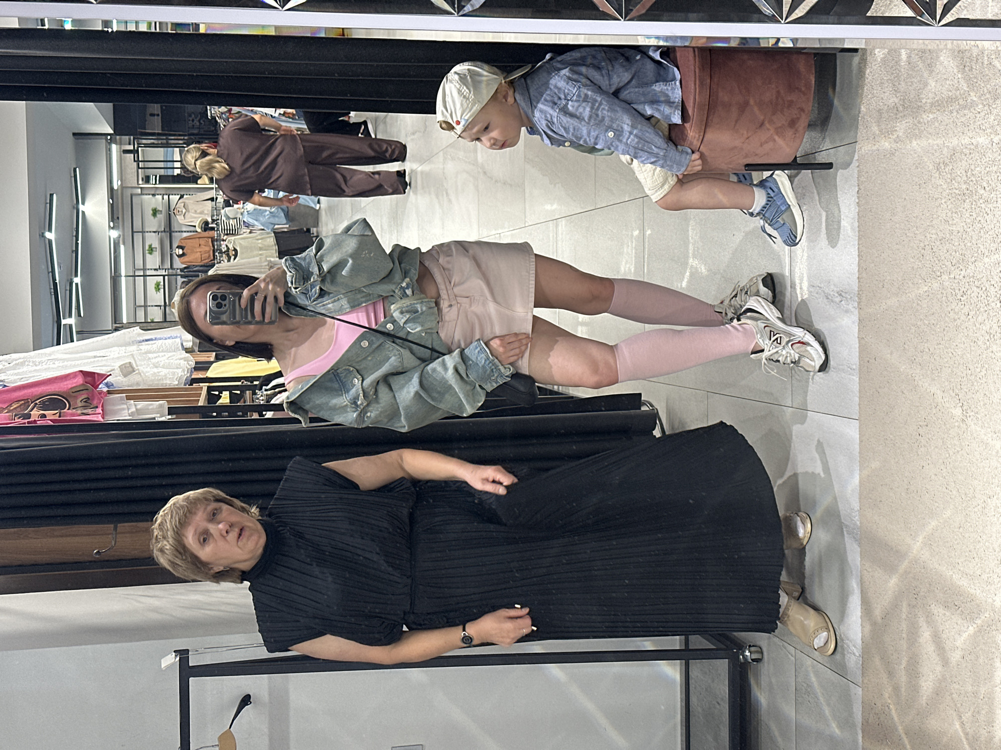 In a store in Votkinsk, Russia, a woman is taking a selfie in a mirror. She is wearing a black pleated dress and light-colored sandals. The mirror also reflects a young boy wearing a blue shirt, beige shorts, and a white baseball cap. He is sitting on a brown stool. In the background, clothing racks and displays of apparel are visible, suggesting this is a retail environment. The lighting appears to be artificial, typical of an indoor commercial space. The woman's expression is neutral, and the boy appears to be looking down. The image is taken from a low angle, capturing the floor and lower parts of the store fixtures.