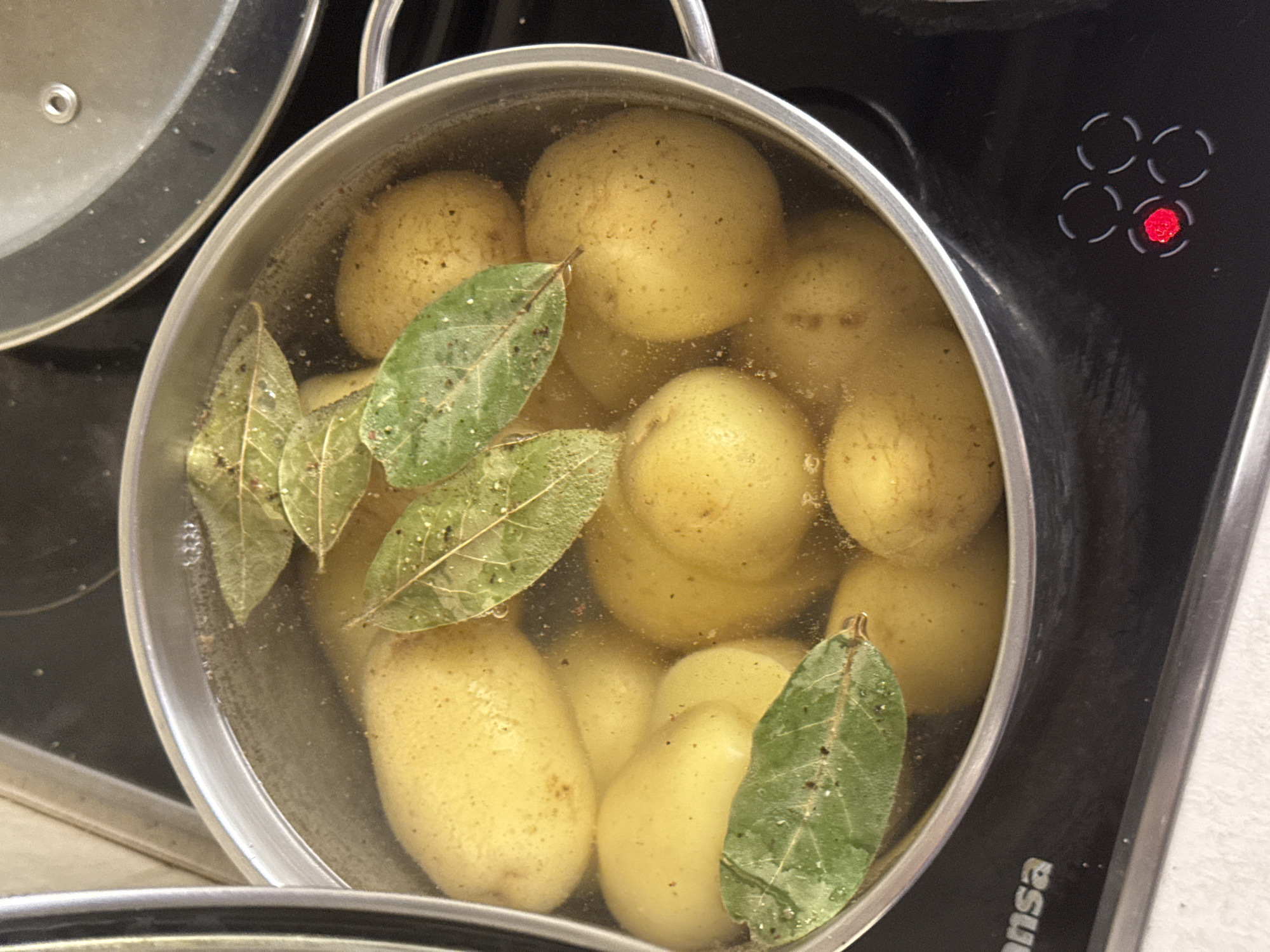 A pot of small, whole potatoes is being boiled on a black stovetop. The potatoes are partially submerged in clear liquid, and several bay leaves are floating on top. The stovetop has indicator lights, one of which is red, suggesting it is currently in use. The pot is a stainless steel saucepan with handles. A second pot with a glass lid is partially visible in the background. The environment appears to be a kitchen. No people are visible in the image, and no specific location in Budva, Montenegro, can be determined from the visual information.