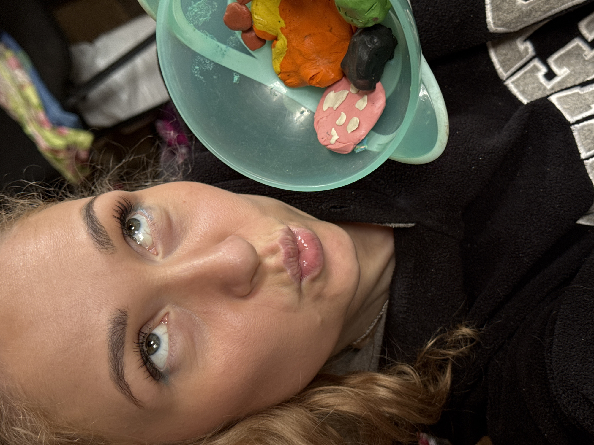 A selfie of a light-skinned woman with blond hair, blue eyes, and pursed lips is displayed. She is wearing a black sweater with white letters. Above her face is a light blue bowl containing multi-colored modeling clay. The background is blurry, with indistinct shapes and colors, but appears to be a cluttered indoor setting. The location, Rosstani, Russia, is not identifiable in the image.