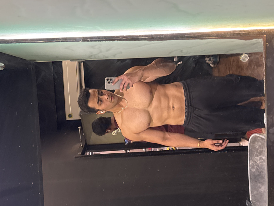 The image displays a male individual capturing a mirror selfie within an indoor setting, identified as a gym or changing room in Kolkata, India. The primary subject is shirtless, exhibiting a muscular physique with visible pectorals, deltoids, and abdominal definition. He wears dark athletic pants or joggers, a gold-colored chain necklace, a white wireless earbud in his right ear, and a gold-colored bracelet on his left wrist. He holds a silver-colored smartphone with his right hand to photograph his reflection. In the background, visible in the mirror's reflection, another individual is partially discernible from behind, wearing a dark red shirt. The environment features dark-colored walls and an air conditioning unit mounted high on the wall. A vertical strip of light is reflected along the right edge of the mirror.