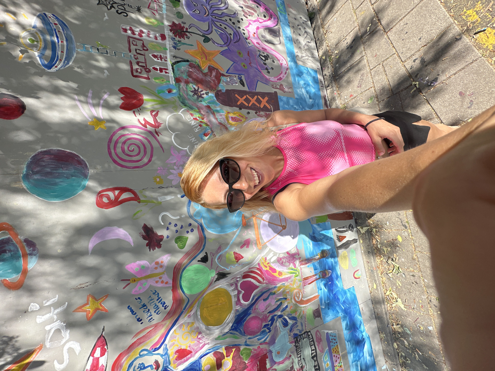 A blonde woman with sunglasses and a bright pink top is taking a selfie in front of a colorful mural on a grey wall. The mural depicts various whimsical drawings, including planets, a UFO, a bicycle, flowers, and an octopus. Some of the drawings appear to be children's artwork. The woman is smiling and holding her arm out to take the photo. She is wearing black shorts. The ground in front of the wall is paved with stone tiles, and there are some fallen leaves on the ground. The scene appears to be outdoors on a sunny day, with dappled shadows from trees falling on the wall and ground. Based on the mural's distinctive style and motifs such as the 'XXX' symbol often associated with Amsterdam, the location is likely in the city of Amsterdam, Netherlands.