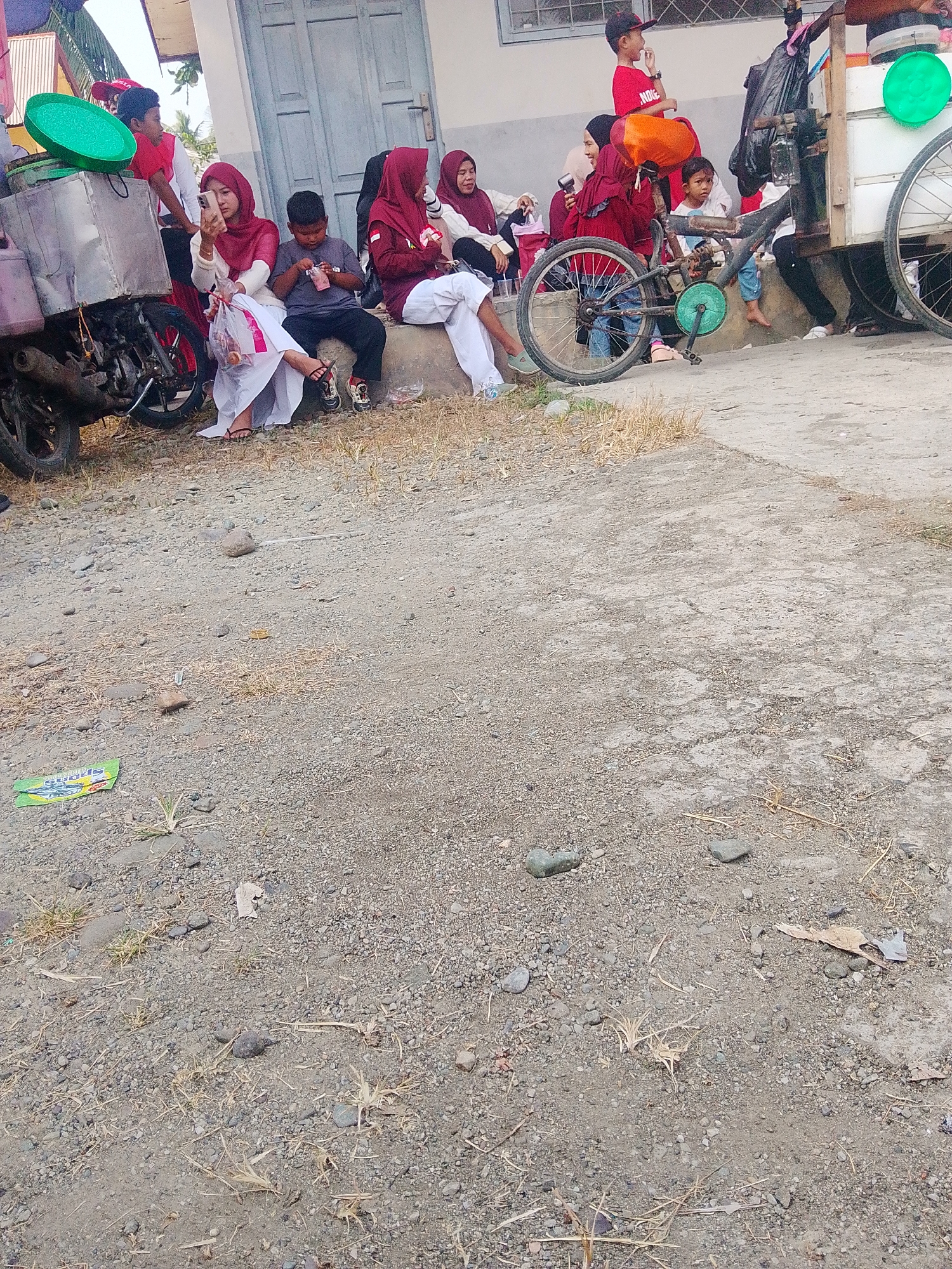 In a street scene in Barabai, Indonesia, a group of children and adults are gathered. A motorcycle with a sidecar, likely used for selling goods, is parked to the left of the frame. The sidecar is a metallic box structure, and on top of it rests a large, bright green circular object. Several individuals, including children in red shirts and adults in hijabs, are seated on a raised concrete curb in front of a building with a gray door and windows. One woman in a red hijab is holding a mobile phone, seemingly taking a photo or video. A child in a dark grey shirt is looking down at something in his hands. A bicycle with a green pedal crank is positioned in front of the seated group, its front wheel partially obscured. The ground is covered with gravel and dry grass. In the background, a building with a light-colored facade is visible.