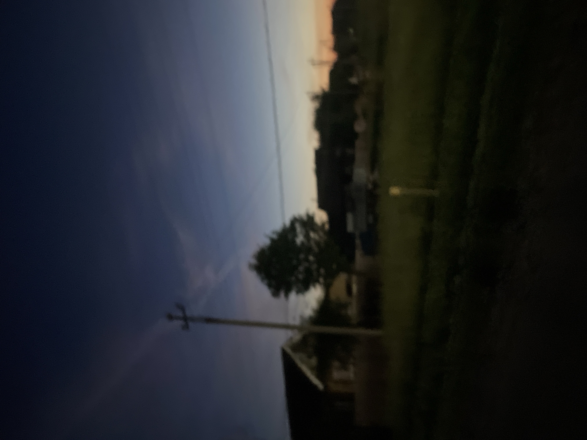 The image depicts a rural scene at dusk in Krasnye Baki, Russia. The sky is a deep blue transitioning to orange and yellow hues near the horizon, suggesting sunset or sunrise. Silhouetted against the sky are several houses with dark roofs and a prominent utility pole in the foreground. A large tree stands between the pole and some of the houses. The ground is covered in grass, appearing dark and somewhat overgrown. A faint light source, possibly a streetlamp, is visible in the middle ground on the left. The overall impression is one of a quiet, undeveloped area. There are no people or animals visible in the image.