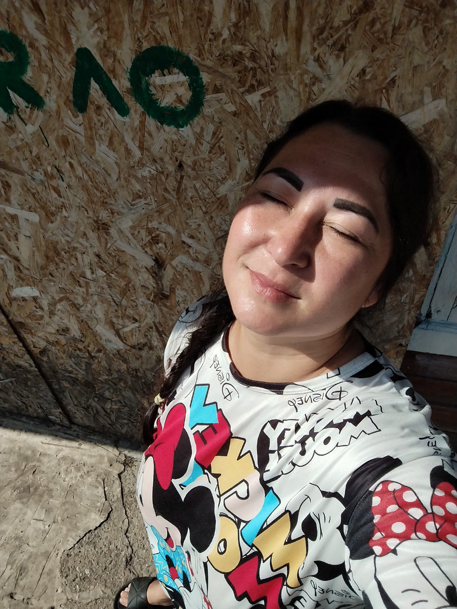A woman is captured in a close-up, high-angle selfie outdoors in Krasny Gulyai, Russia. She is standing in front of a wall made of oriented strand board (OSB) with green painted letters that appear to spell "RAO." The wall is rough and textured, showing the wood chips embedded within it. The ground beneath her is concrete, with visible cracks. Sunlight is casting a strong shadow from her left side, highlighting the contours of her face. She has her eyes closed, her head tilted back slightly, and a subtle smile on her lips, suggesting she is enjoying the sun. Her dark hair is pulled back, and a braid is visible over her shoulder. She is wearing a white long-sleeved shirt adorned with a graphic print featuring characters and text, possibly from Disney, including Minnie Mouse. Her left foot, wearing a black sandal, is visible at the bottom of the frame. The scene appears to be in a somewhat unkempt outdoor area, possibly a construction site or an abandoned space.