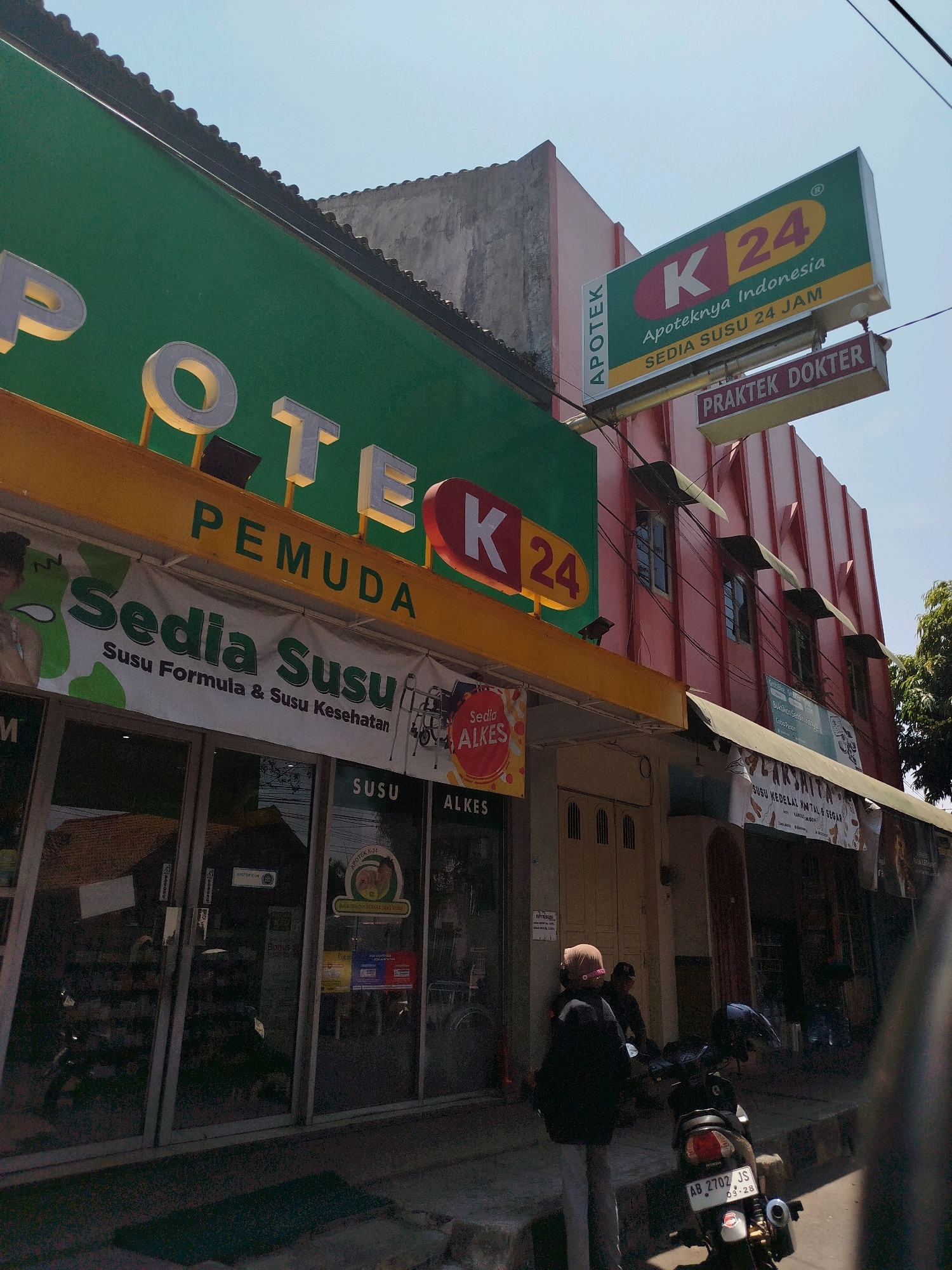 The image captures a street scene in Kebumen, Indonesia, focusing on a K24 Apotek building. The building features prominent signage. A large green sign with "POTE K" is visible on the left, suggesting the name "APOTEK PEMUDA K24". To the right, a rectangular sign with a green background and yellow lettering displays "K 24 Apoteknya Indonesia SEDIA SUSU 24 JAM". Below this, a smaller sign reads "PRAKTEK DOKTER". The storefront appears to be an apothecary, with various products advertised on banners, including "Sedia Susu" (Milk is available), indicating "Susu Formula & Susu Kesehatan" (Formula Milk & Health Milk). Further down the street, another establishment has signage that includes "LAKSMITA" and "SUSU KEDELAI HALAL & SEGAR" (Halal & Fresh Soy Milk). Two individuals are seen near a motorcycle parked on the sidewalk, with one person wearing a hijab and another person behind them. The environment is sunny with a clear blue sky, and the architecture suggests a typical urban setting in Indonesia. The motorcycle's license plate reads "AB 2702 JS".