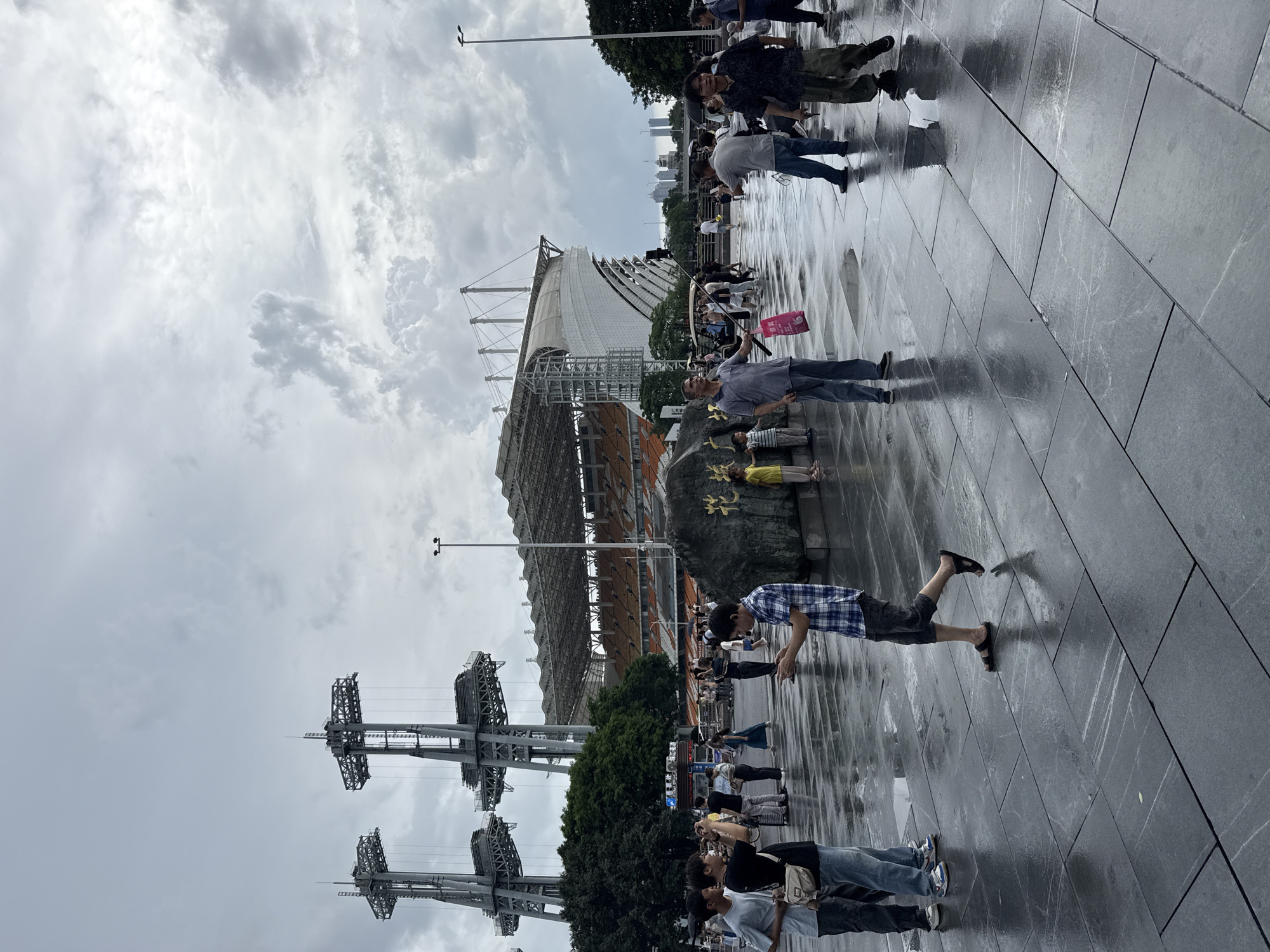 The image shows a wide-angle view of a paved plaza in front of a large stadium with a distinctive white, curved facade and an orange-seated grandstand. Two tall, industrial-looking towers with complex metal structures are prominent in the background, to the left of the stadium. The plaza is wet, indicating recent rain, and reflections of the sky and surrounding elements are visible on the dark, glossy paving stones. Several people are scattered across the plaza, some walking, some standing, and some appearing to be taking photographs or looking at their phones. In the foreground, on the right side of the image, is a large, dark rock with some golden text inscribed on it. The sky is overcast with dramatic, heavy clouds. The overall scene suggests a public space outside a major sporting or event venue in a city. The location is Tianhe District, China.