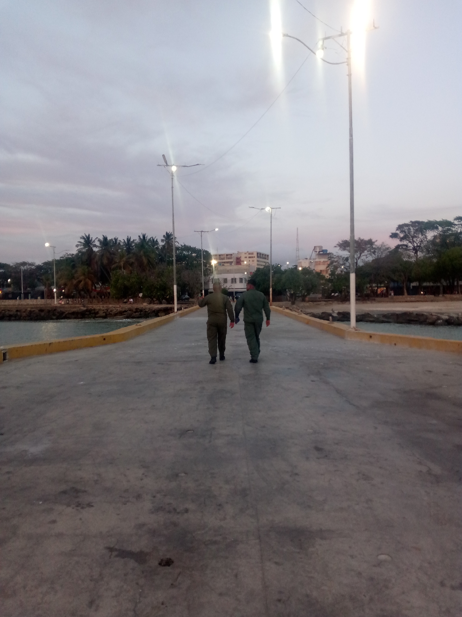 Two people in military-style uniforms walk hand-in-hand down a long, concrete pier towards a shoreline. Streetlights line the pier, and buildings and trees are visible along the shore. The location appears to be a city waterfront, possibly in Unknown. The sky is overcast.