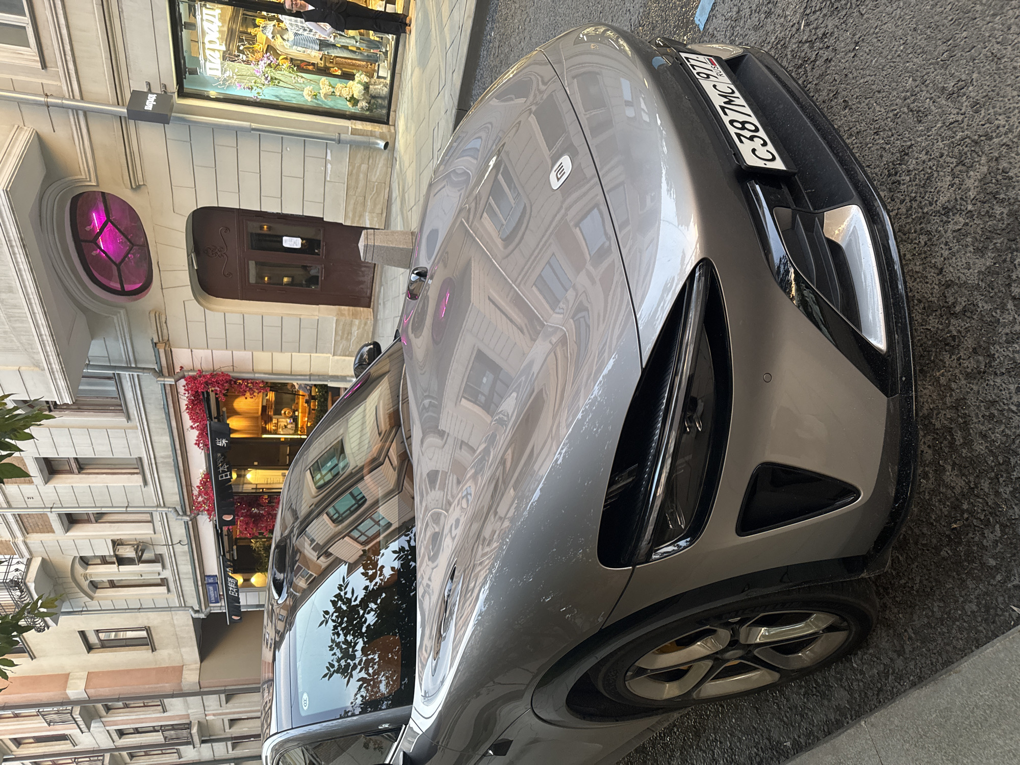 A grey Xiaomi SU7 electric vehicle is parked on a cobblestone street in Moscow, Russia. The car is angled towards the camera, with its front end prominently displayed. The vehicle has a sleek design with sharp lines and distinctive LED headlights. A license plate reading "C387MC 977 RUS" is visible on the front bumper. In the background, a multi-story building features a decorative oval window with a pink interior light and an ornate brown door. A shop window with mannequins dressed in clothing and floral displays is also visible. The street is lined with trees and buildings, creating an urban cityscape. A person in a dark suit is standing outside the shop. The overall scene suggests a sunny day in a European city.