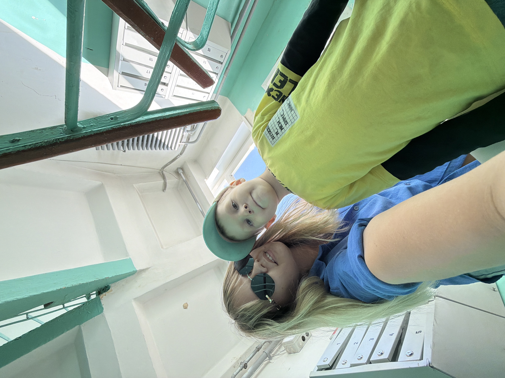 Two people, presumably a parent and child, are positioned in the stairwell of a building. The adult is wearing sunglasses and a blue shirt, while the child is wearing a green cap and a yellow long-sleeved shirt. The stairwell features mint green paint on the railings and walls. Mailboxes are visible with numbers 13, 14, 15, 17, and 18. The setting appears to be an apartment building. The shirt worn by the child has the words "NEW GAME SCORE" written on it. The image was taken from below. The location is Chaikovsky, Russia.