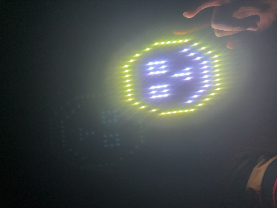 A circular LED display shows the number 24, surrounded by a yellow border of lights. A hand is positioned above the display. A second, fainter LED display, also displaying the number 24, is visible in the background. The scene is dark, with a hazy, out-of-focus quality, possibly due to low light conditions or a camera lens effect. The background is mostly obscured by darkness. A partial view of what seems to be clothing is visible on the right.