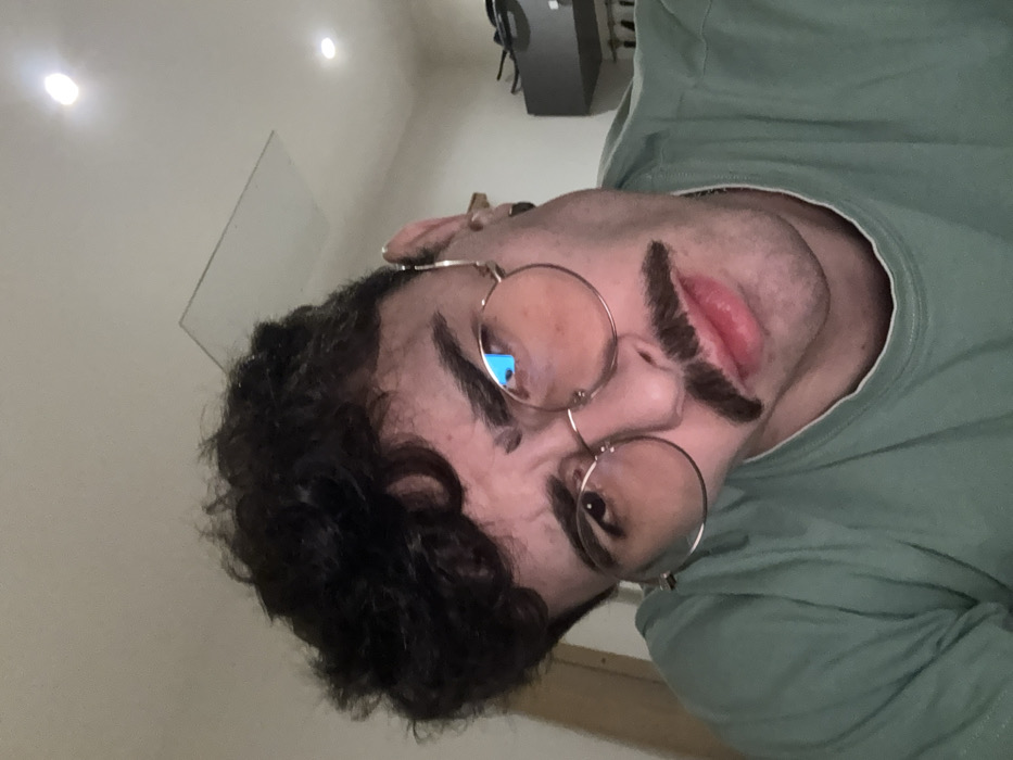 A person with dark curly hair, round glasses, and a thick, fake mustache is angled in the frame. The person wears a light green shirt. Above the person, three ceiling lights are visible, along with a white board and a gray cabinet. The picture is taken from a low angle, making the person's face the focal point.