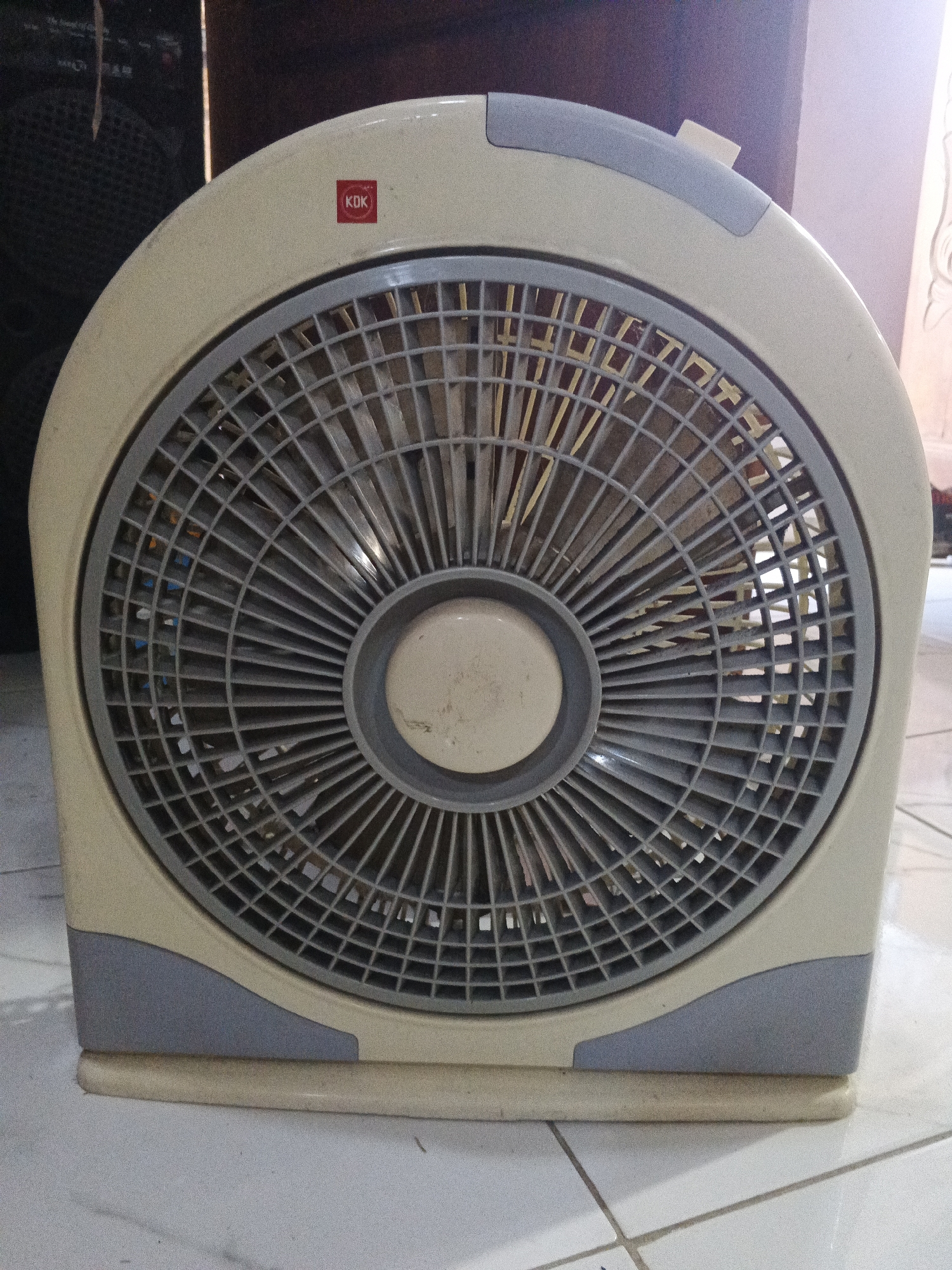 The image shows a close-up, eye-level view of a beige box fan with a gray grille and accent pieces. The fan is positioned on a light-colored tiled floor. The fan has a circular grille with many thin, vertical slats. In the center of the grille is a circular, beige hub. A small, red sticker with the white text "KDK" is visible on the upper right portion of the fan's casing. The background is out of focus, but appears to include dark objects, possibly speakers or furniture, and a light-colored wall or curtain. The lighting suggests an indoor environment. There are no people or animals visible in the image. The overall impression is of a functional household appliance. The specific location in Kemangsen, Indonesia, cannot be determined from the visual information present in the image.