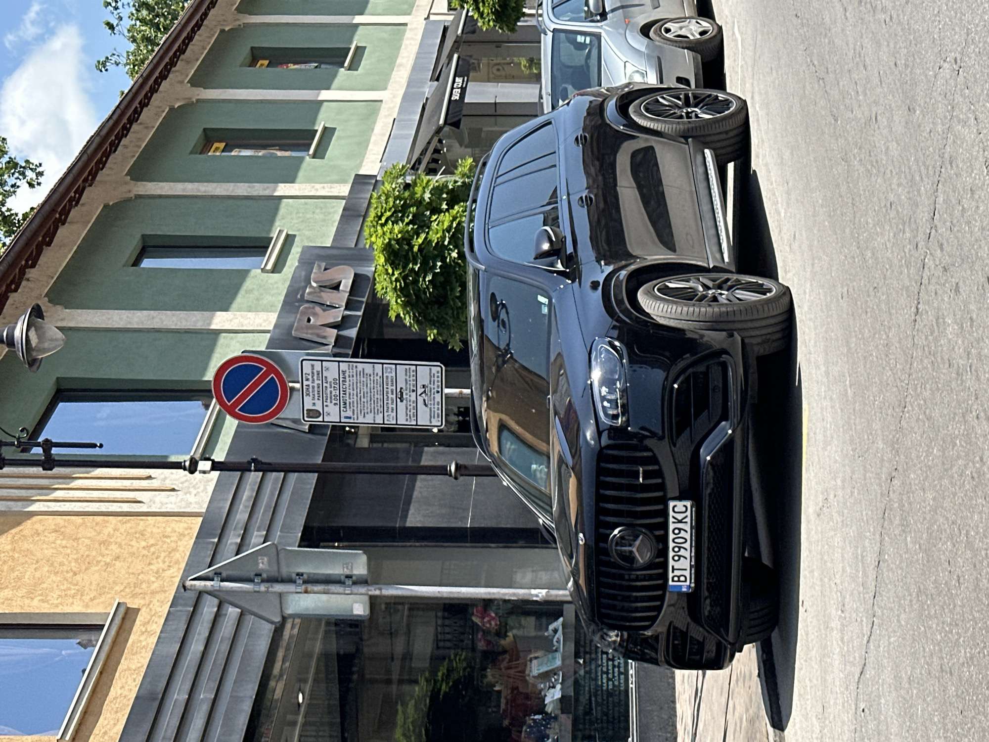 The image captures a street scene in Veliko Tarnovo, Bulgaria, featuring a black Mercedes-Benz SUV parked in front of a building. The car's license plate reads "BT 9909 KC." Next to the car is a "no parking" sign and a sign indicating a paid parking zone, operational on weekdays from 9:00 to 17:00, labeled as Zone No. 2. The building behind the car has a sign with the word "RIVIS".