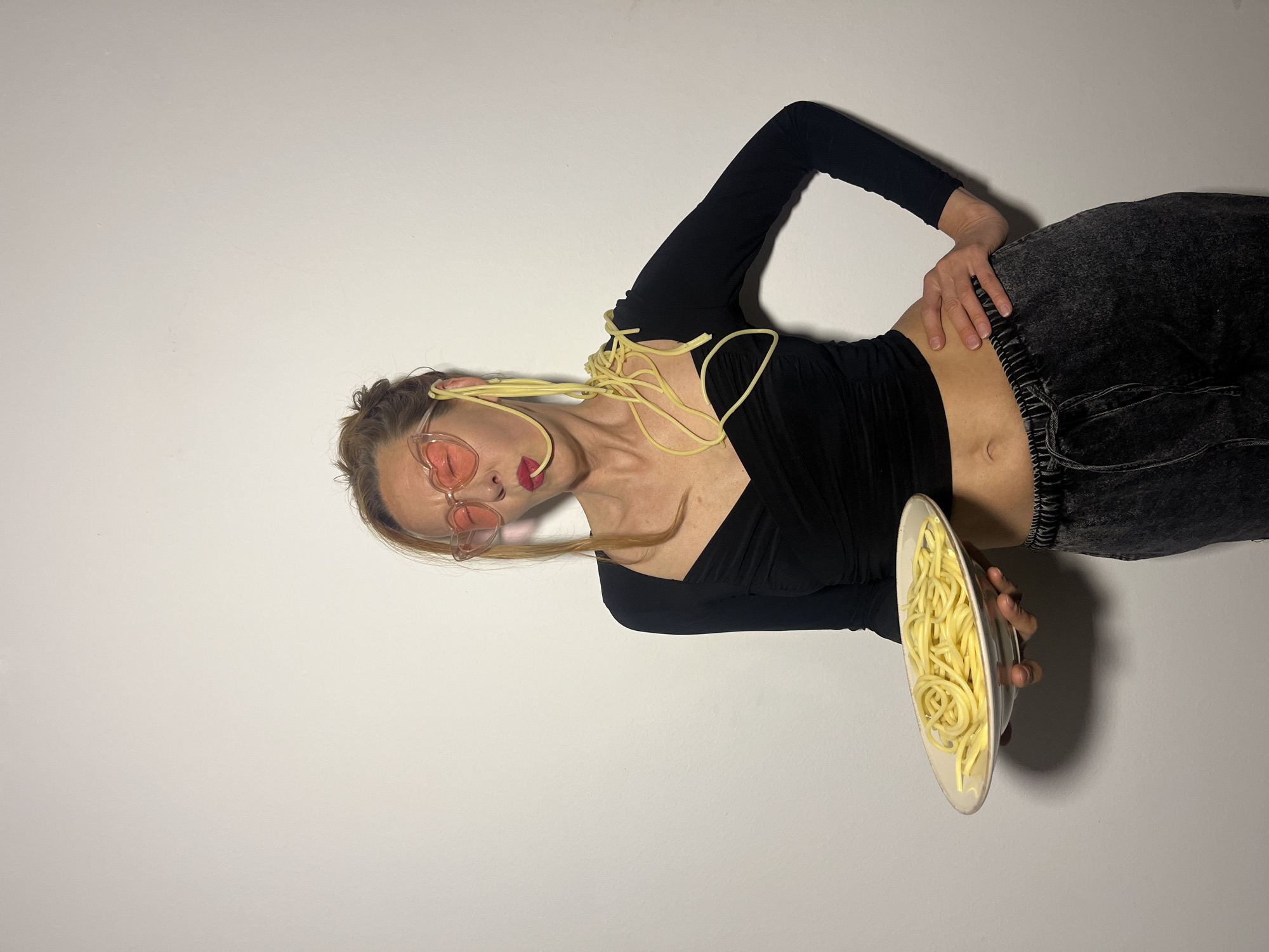 The image displays an adult female subject standing against a plain, light-colored wall. She has light brown hair, pulled back, and wears translucent pink-red heart-shaped eyeglasses along with red lipstick. Her attire includes a black, long-sleeved, ruched crop top that exposes her midriff, and dark, acid-wash-style sweatpants with a visible drawstring. The subject holds a white plate filled with cooked spaghetti in her left hand, extended forward. Additional strands of cooked spaghetti are draped over her right ear, across her right cheek, neck, and shoulder, with some resting on her chest. A single strand of spaghetti protrudes from her mouth. Her right hand is positioned on her right hip. The scene is located in Budva, Montenegro.