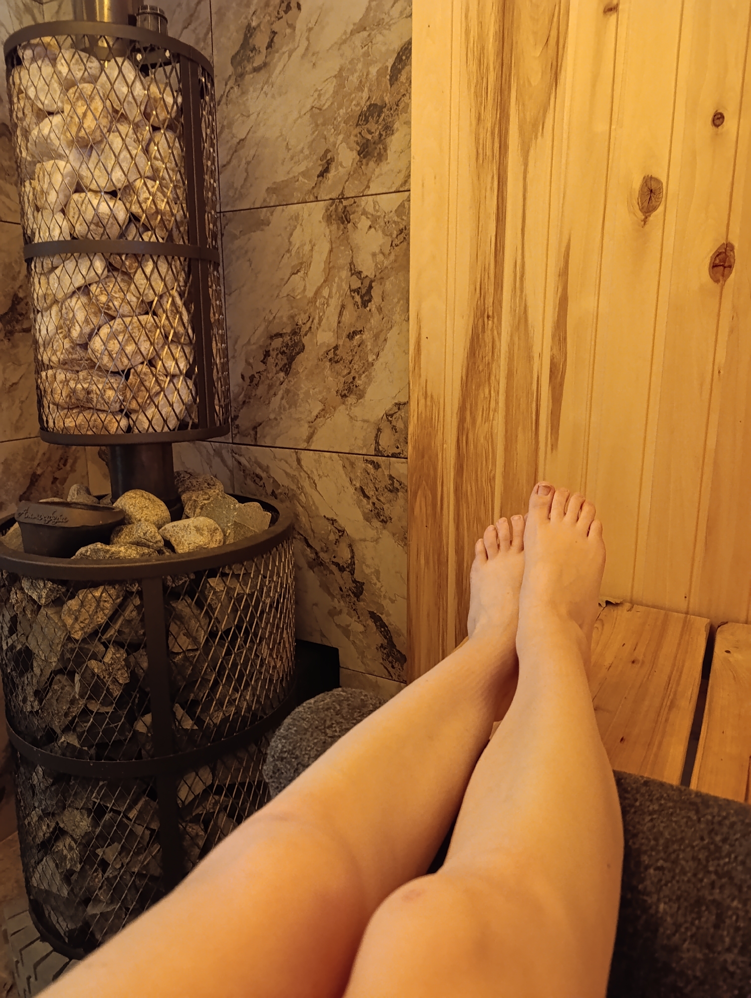 The image depicts a person's legs and feet resting inside what appears to be a sauna. The person is lying on a wooden bench. Behind the person is a wood-paneled wall and a tile wall. To the left, there is a metal sauna stove filled with stones. Based on the location provided, this scene is likely taking place in a sauna in Chaikovsky, Russia.