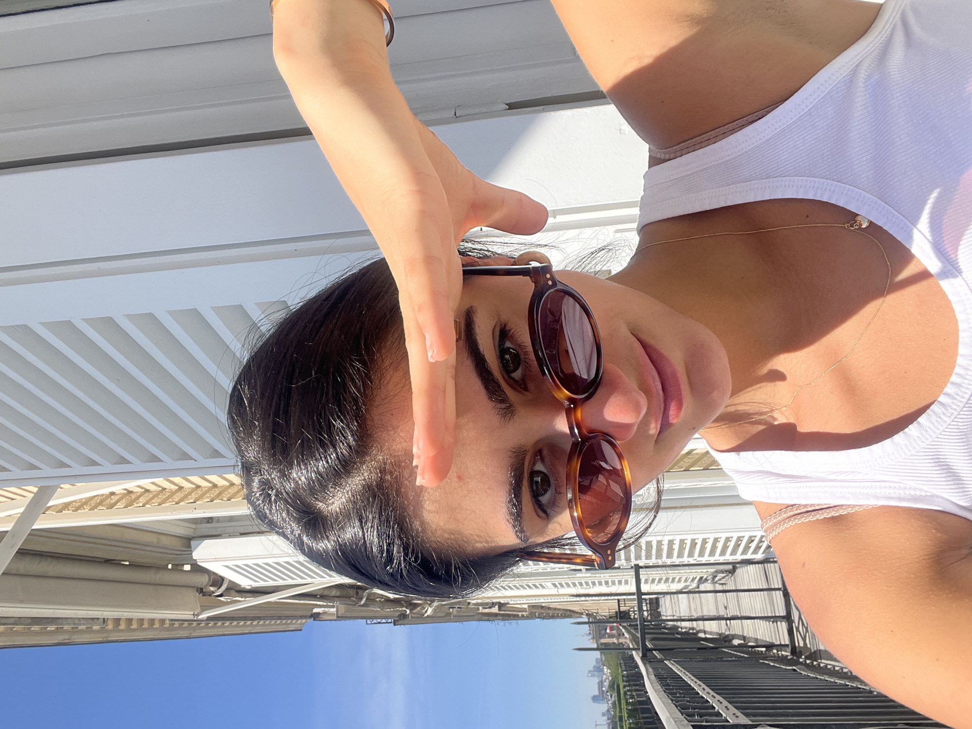 A young woman is captured in a close-up, upward-angled shot outdoors, possibly on a rooftop or balcony in Paris, France. She has dark hair, styled with visible braids or cornrows near her forehead, and is wearing sunglasses with tortoise-shell frames. Her right hand is raised, partially shading her face from the bright sun, with a subtle golden bracelet visible on her wrist. She is looking towards the camera with a relaxed expression, her lips slightly parted.
She is dressed in a simple white ribbed tank top, and a delicate gold chain necklace with a small pendant is visible on her neck and chest. The background features a distinctive white slatted structure, likely an awning, pergola, or architectural element, indicating an outdoor setting. Beyond this, a clear blue sky extends, with a distant cityscape visible on the horizon, confirming an urban environment. Strong sunlight casts distinct shadows, suggesting it is daytime, likely late morning or afternoon. There is no visible text in the image.
