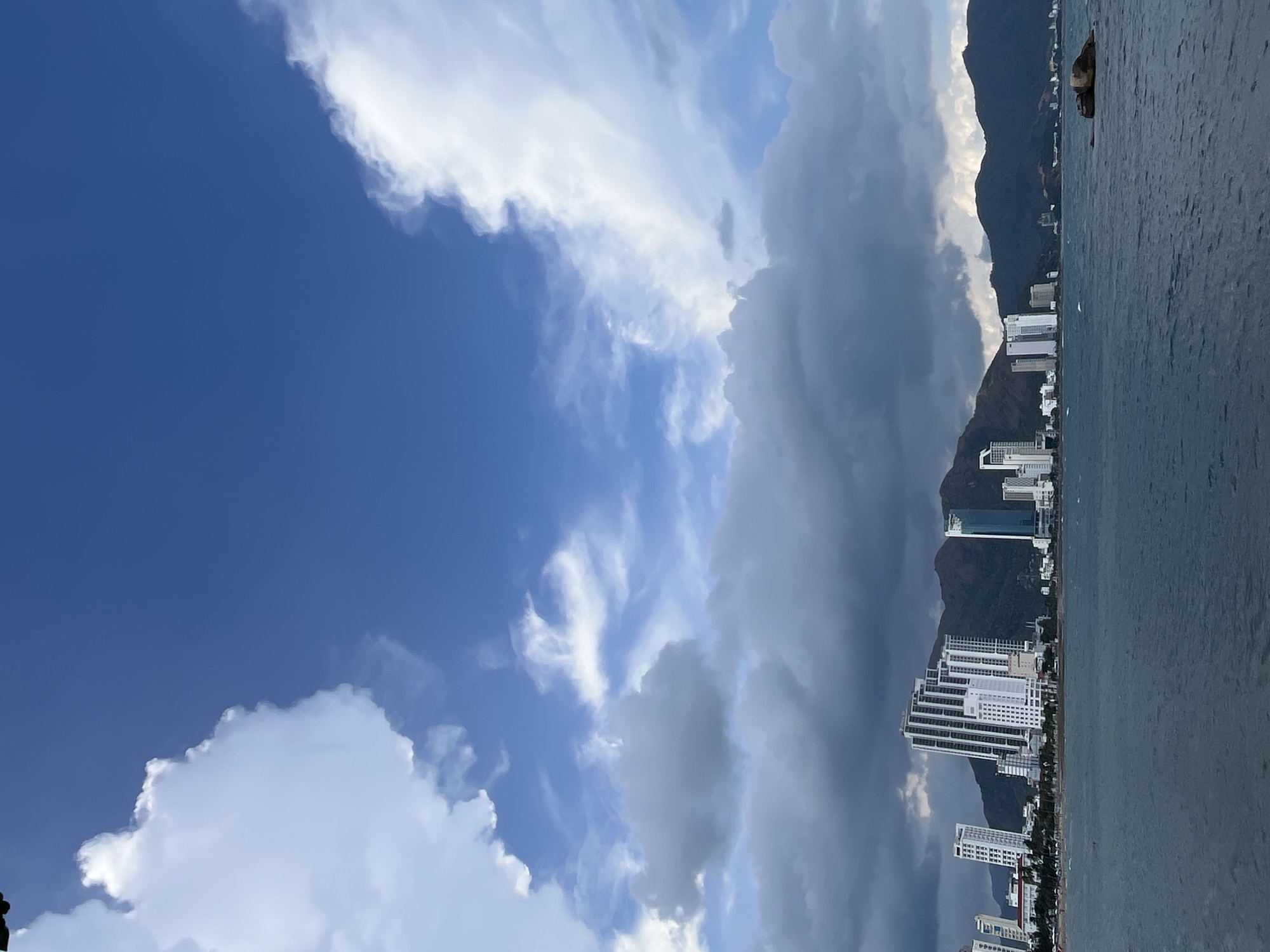 The image shows a coastline view in Bac Nha Trang Ward, Vietnam, with a contrast of weather conditions. The left side features a clear blue sky with white cumulus clouds, while the right side displays dark storm clouds hovering over a mountainous area adjacent to the coastline. Several tall, modern buildings are situated along the shore, backed by the mountains. The sea is visible in the foreground, reflecting the light and colors of the sky. There are no people or visible actions or interactions in the scene.