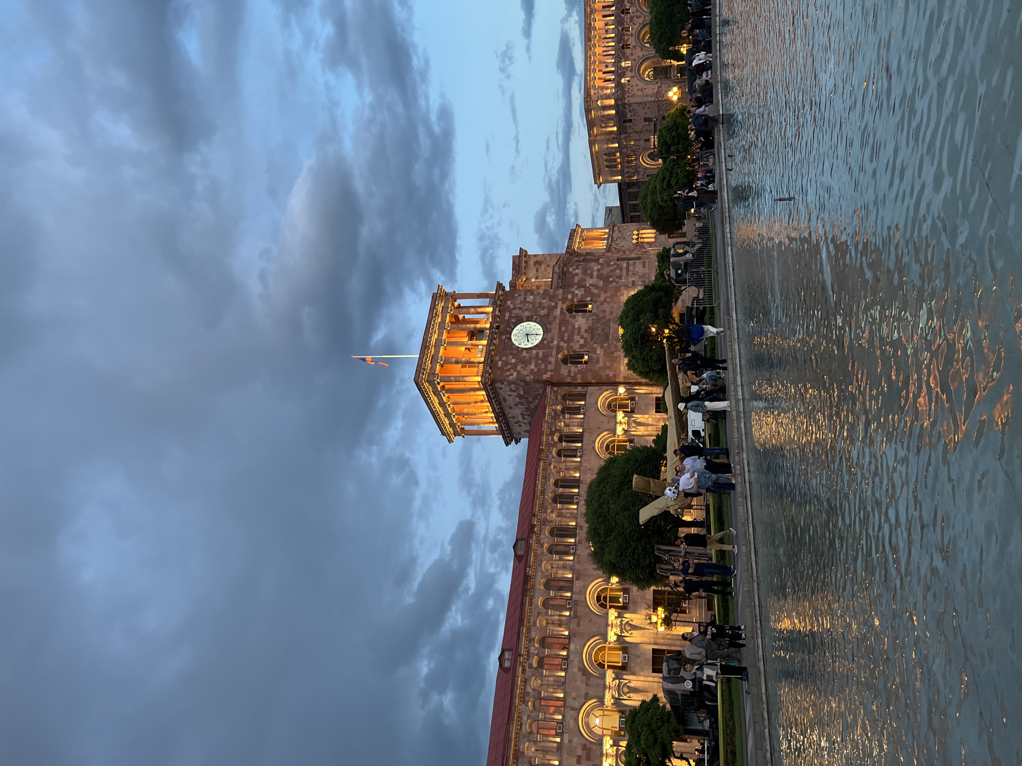 The image depicts a view of Republic Square in Yerevan, Armenia, at dusk. The building with a clock tower, likely the Government House, is prominently featured, illuminated with warm artificial light contrasting with the darkening sky. A body of water, possibly a fountain or reflecting pool, occupies the right side of the frame, its surface reflecting the lights from the building. People are present along the edge of the water feature, some seated at what appear to be outdoor cafes or seating areas. Trees are interspersed between the buildings and the seating areas. The sky is overcast with heavy clouds. An Armenian flag is visible atop the clock tower building. The overall scene presents a public space being used by people in the evening hours.