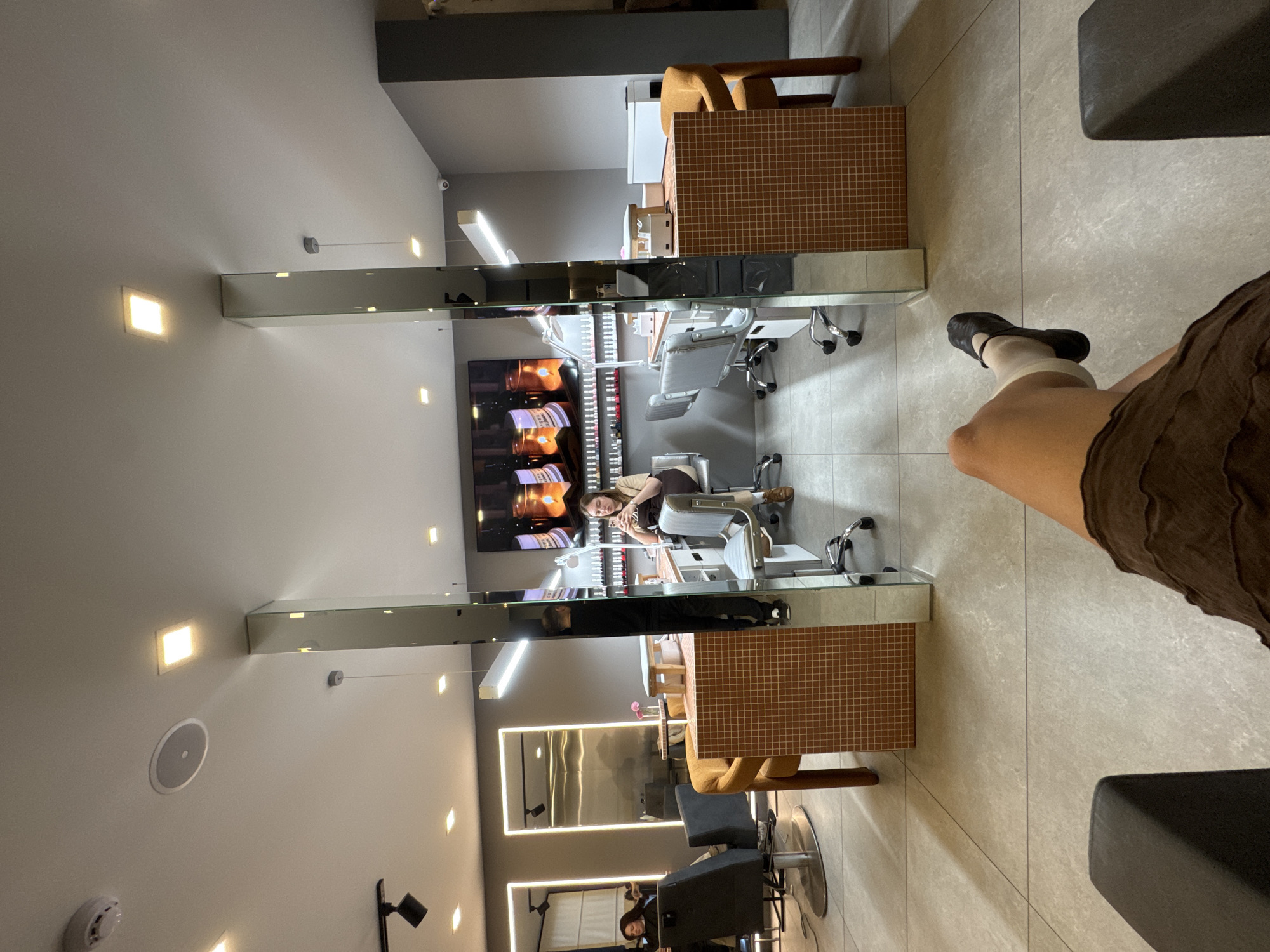 A first-person, indoor shot depicting a leg clad in a brown, textured garment and a black shoe with a light-colored sock. The setting appears to be a beauty salon in Tbilisi, Georgia, judging by the arrangement and style of salon furnishings.
The interior of the salon is visible through a mirror, with a manicurist attending to a client. A grid-patterned countertop and salon chairs are also in view. The reflection includes other salon workers and clients, suggesting a busy environment. Overhead lighting is embedded in the ceiling, and the room's aesthetics include mirrored surfaces and modern chairs.