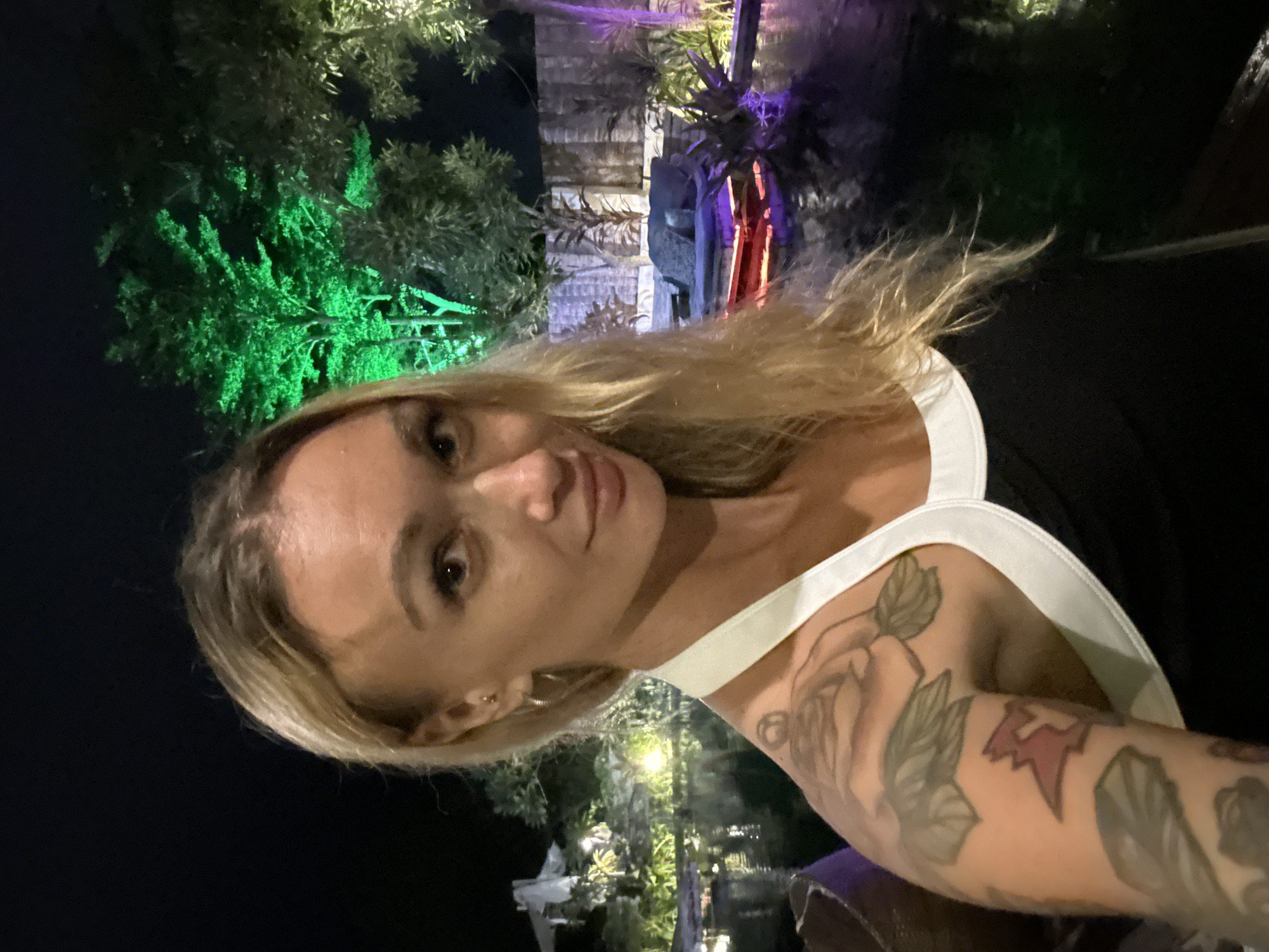 A blonde woman with tattoos on her left arm takes a selfie at night. She is wearing a black top with a white trim. The background shows a body of water, lush green trees illuminated by green lights, and a lounge area with purple lighting. This appears to be an outdoor setting at a resort or a similar establishment in Mũi Né, Vietnam, a coastal resort town known for its beaches and sand dunes.