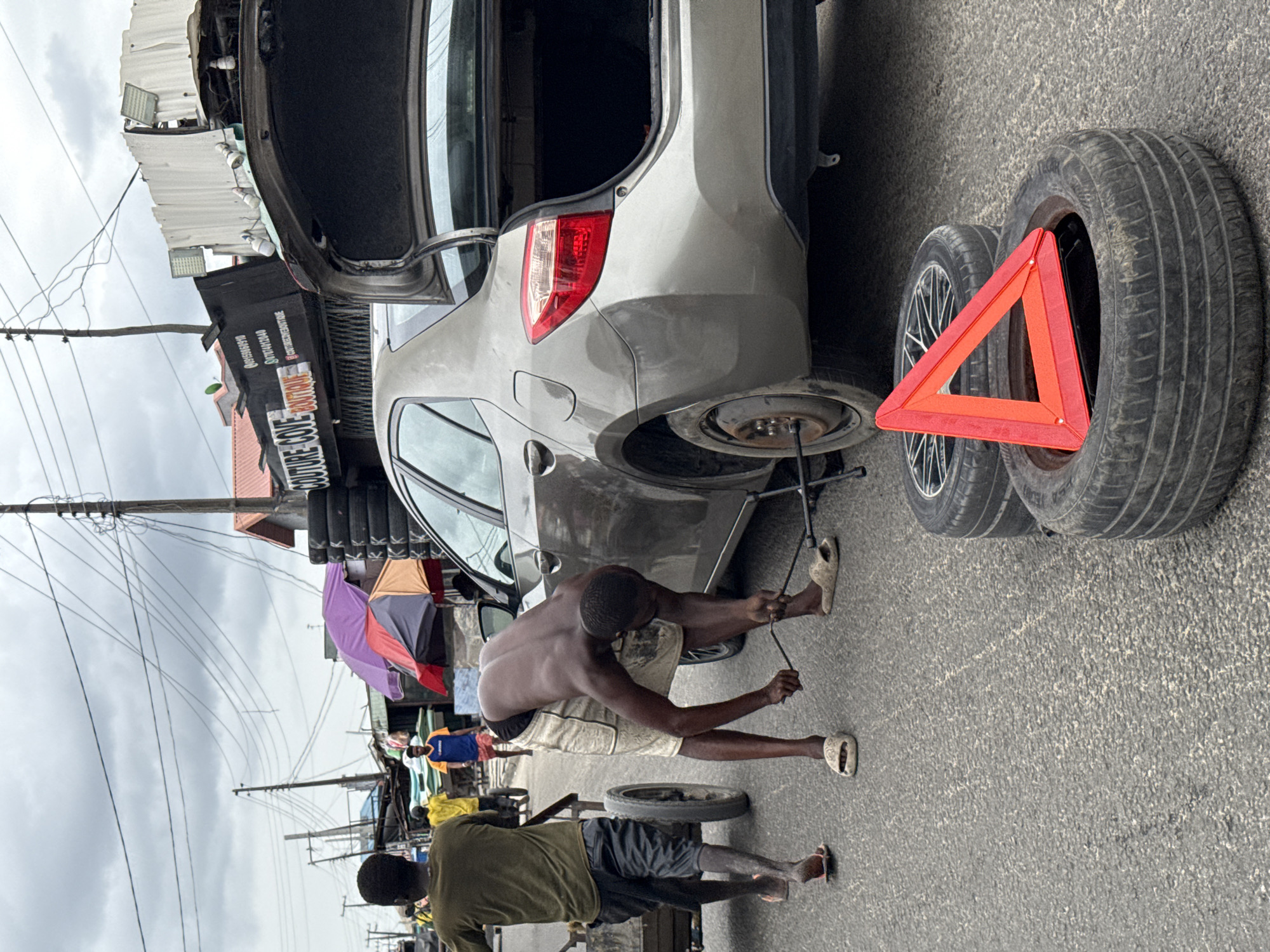A man is changing a tire on a silver sedan with the trunk open on a street in Ado, Nigeria. He is using a lug wrench. Two tires with a red warning triangle are leaning against the car. A second man is nearby. Overhead power lines, market umbrellas, and commercial signs are visible in the background. A building is labeled "COUTURE COLE".