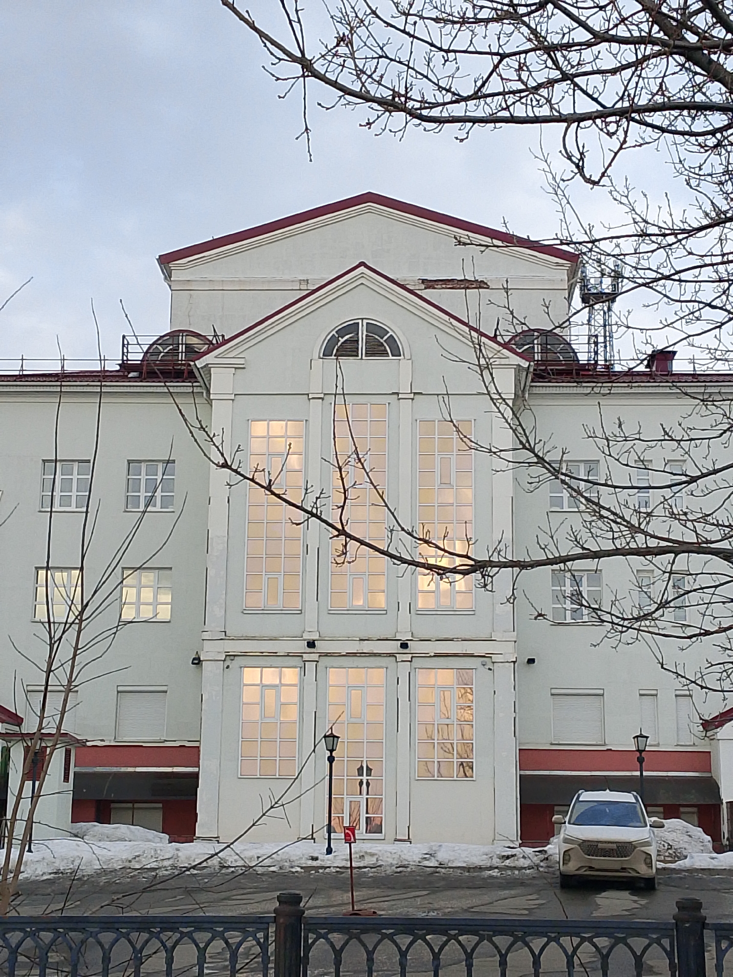 The image captures a frontal view of a white building with a red roof, likely located in Izhevsk, Russia. The building features tall, multi-paned windows that glow with interior light. Bare tree branches obscure portions of the façade. A car is parked in front of the building, partially obscured by snow piles. A black iron fence runs along the bottom of the image, partially obscuring the foreground. The sky is overcast. Architectural details include round windows on the roof and a triangular pediment over the central window bay. There are street lamps.