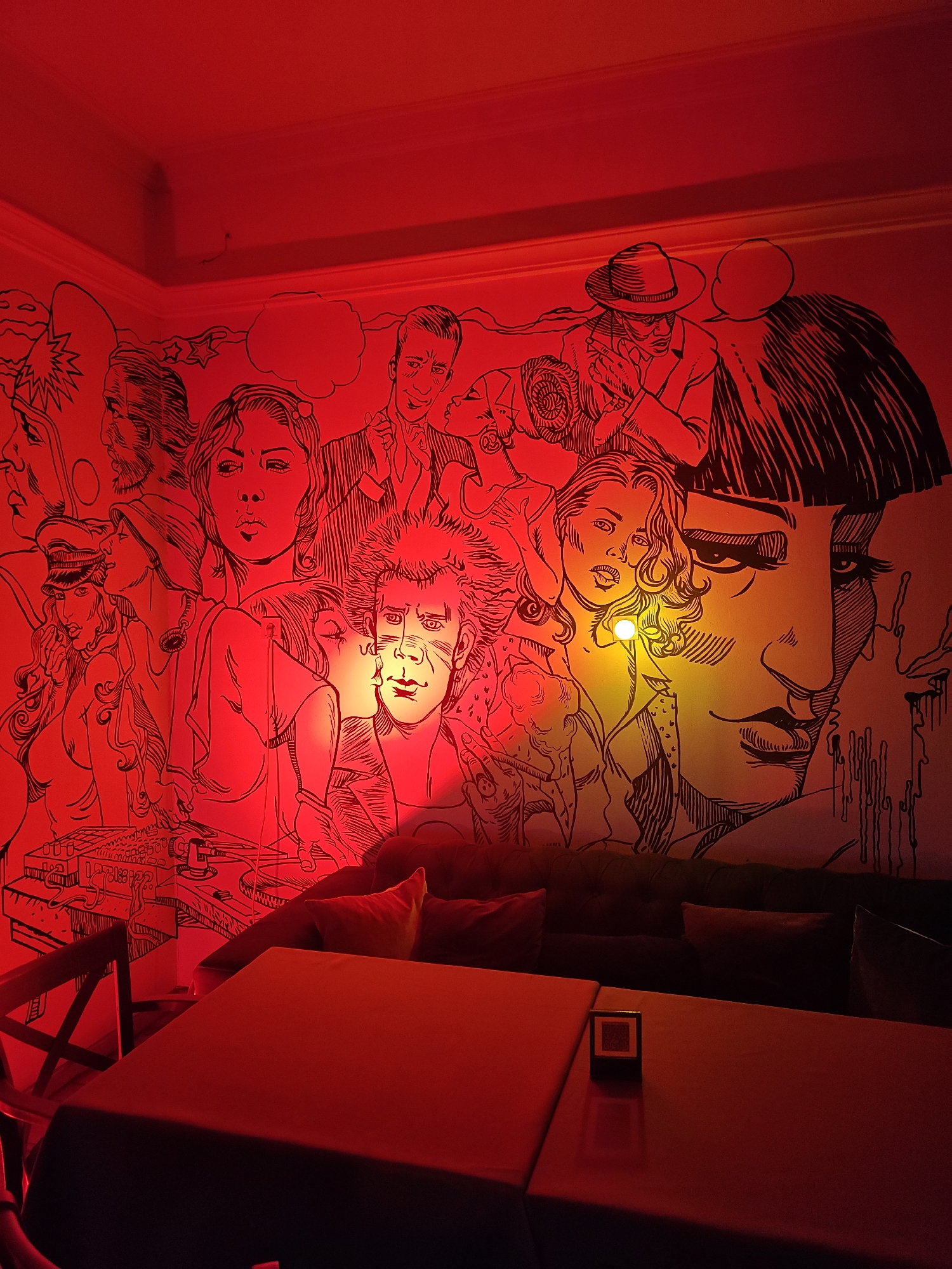 The image shows a room with a large mural on the wall, featuring various figures in a black and white line art style. The lighting in the room is primarily red, giving the scene a reddish hue. The mural includes faces of people, some of whom are partially obscured. The foreground consists of a table with a black frame placed on it, and a couch. The location is Unknown, Unknown.