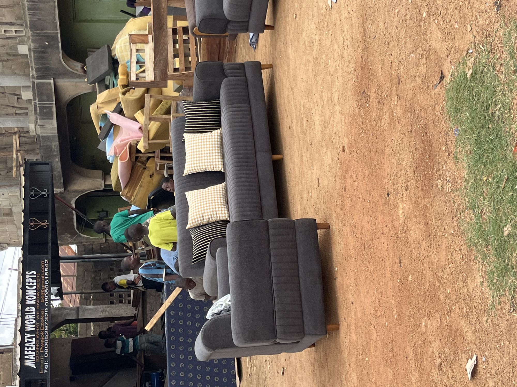 In Bukuru, Nigeria, an outdoor scene depicts a furniture shop with a banner reading "MAFEAZY WORLD KONCEPTS" prominently displayed. The shop appears to be an open-air establishment with various furniture pieces, including a dark grey sectional sofa with patterned cushions, visible in the foreground. To the left, a matching single-seater armchair is positioned. Behind the furniture, stacks of foam and upholstery materials are piled high, suggesting ongoing furniture production or renovation. Several individuals, appearing to be children and young adults, are present in the background, some engaged in activities related to the shop. One individual in a green shirt stands near the stacked materials, while others in casual attire are visible amongst the furniture. The ground is covered in reddish-brown soil, indicative of the Nigerian landscape. The overall environment suggests a bustling commercial area with a focus on furniture sales and manufacturing.