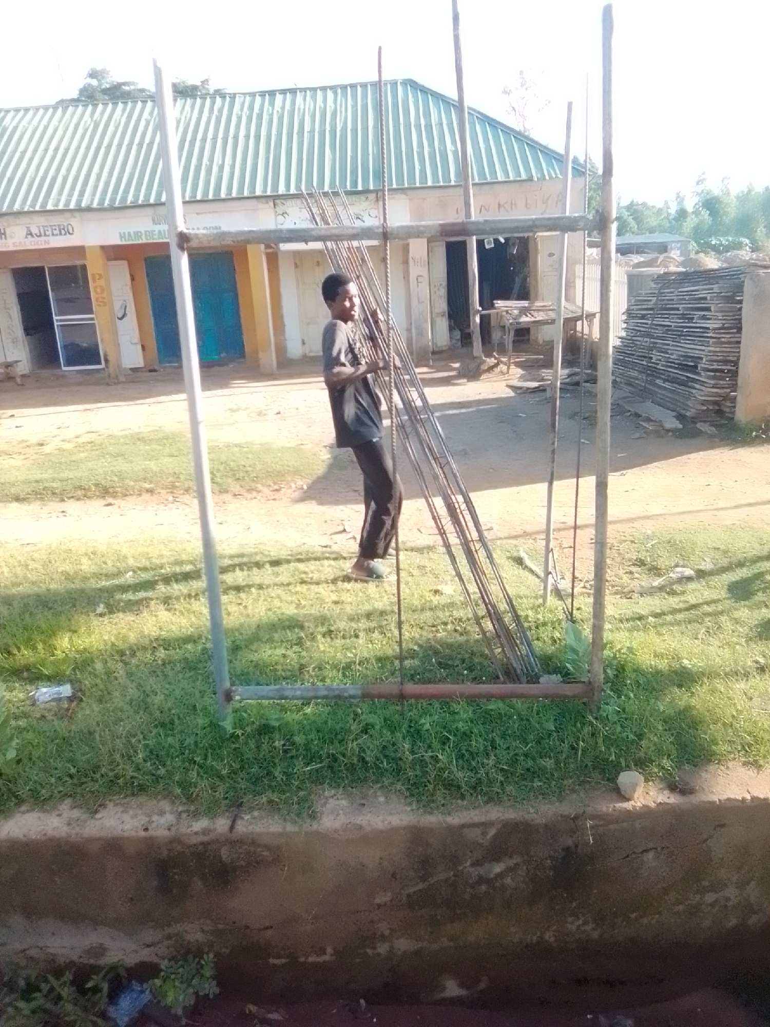 A young male, wearing a dark t-shirt and dark pants with sandals, stands outdoors in Liji Hill, Nigeria, holding a bundle of metal rebar. He is positioned within a frame constructed from metal pipes and wooden poles, suggesting ongoing construction or scaffolding. Behind him, a row of shops with green corrugated roofs is visible. One shop has a sign that reads "HAIR BEAUTY SALOON." Another sign, partially visible, says "AJEEBO." The ground is a mix of dirt and grass. To the right of the shops, a stack of what appears to be lumber or wooden planks is piled high. The overall scene depicts a commercial area undergoing some form of development or renovation.