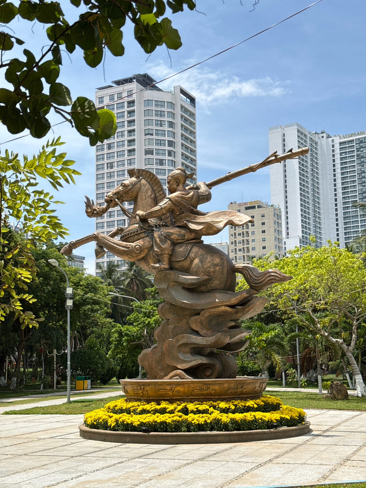 A bronze statue of a warrior on horseback, holding a spear, is prominently displayed in a park in Nha Trang, Vietnam. The statue is set on a decorative base surrounded by yellow flowers. In the background, tall, modern white buildings rise against a clear blue sky with scattered clouds. Lush green trees and palm trees surround the statue, indicating a tropical or subtropical environment. The ground in front of the statue is paved with light-colored square tiles arranged in a grid pattern. The overall scene suggests a public space, possibly a tourist attraction or a landmark within the city of Nha Trang, known for its beaches and resorts. The sunlight indicates it is daytime.