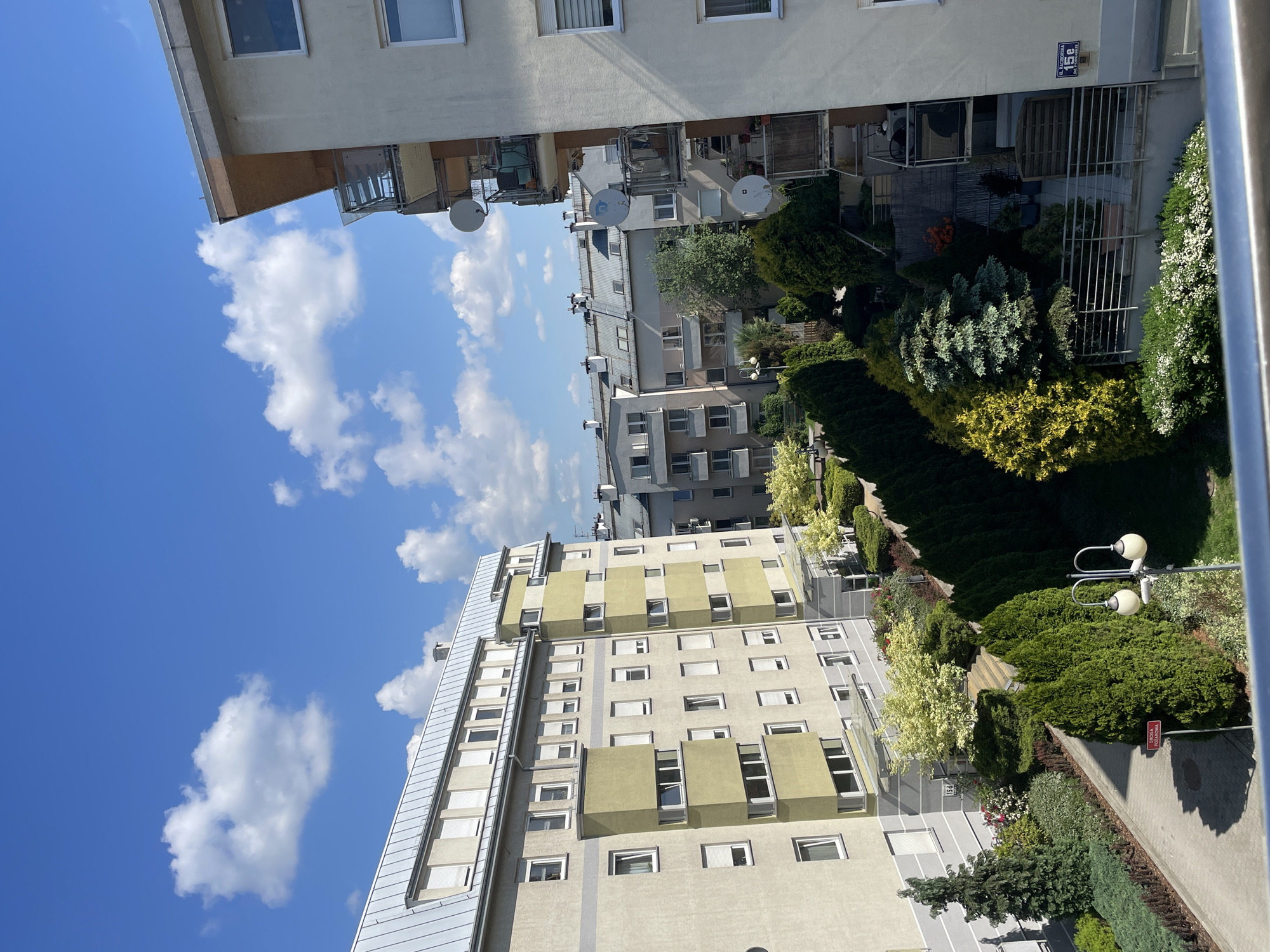 The image shows an urban scene, likely in an apartment complex, during a bright, sunny day. The sky is blue with scattered white clouds. Several multi-story buildings are visible, featuring a mix of architectural styles. One building has a beige facade with protruding, olive green sections around some windows. Another building has satellite dishes mounted on its balconies.
A courtyard area between the buildings is filled with trees and shrubs of varying shapes and colors, primarily green. There's a walkway visible and a few lampposts. A small sign with the number "15c" is visible on one of the buildings. The location appears to be in Unknown, Unknown, based on the signage. There are no people visible in the image.
