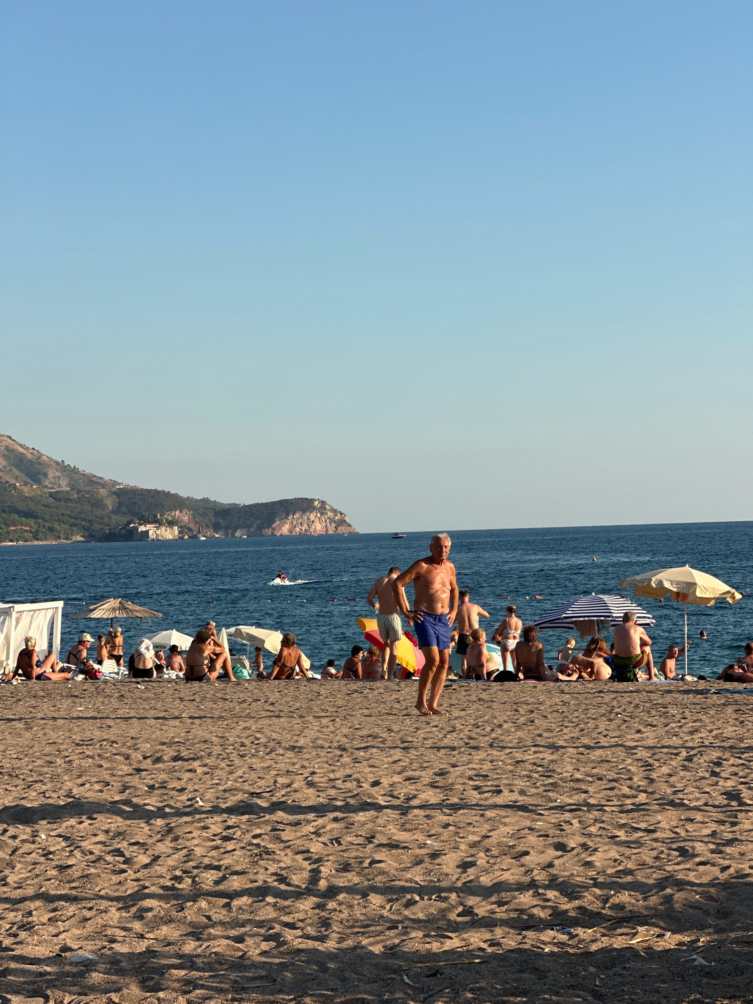 A wide-angle, eye-level shot captures a crowded beach scene on a sunny day at the city of Boreti, Montenegro. The sand-covered beach is populated with numerous sunbathers lounging under umbrellas or directly on the sand, their bodies mostly visible from the torso up as they sit or recline. In the mid-ground, a man in blue swim trunks stands facing left, hands on his hips, his torso bare and aged. The sea occupies the background, its surface calm and blue, with a distant coastline featuring green, tree-covered hills and some buildings. A jet ski is visible on the water, leaving a white wake. The sky is a clear, pale blue, transitioning to a lighter shade near the horizon.