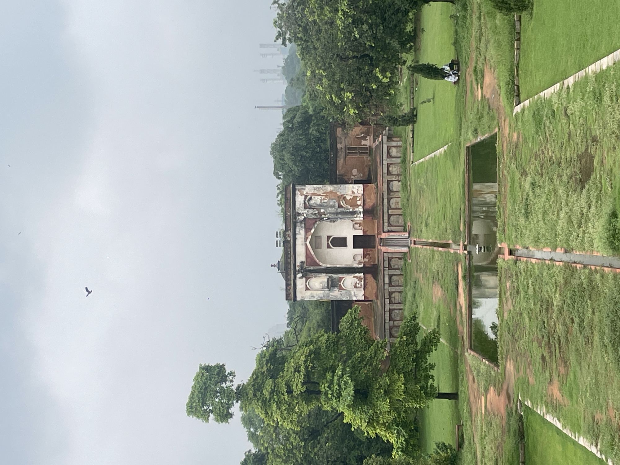 The image depicts a frontal view of a dilapidated building with arched doorways and windows, likely a historical monument in Delhi, India. The building exhibits signs of age and wear, with chipped paint and weathered facades. In front of the building, a symmetrical garden layout is visible, featuring a central pathway lined with water channels and a rectangular pond. Lush green grass covers the ground, interspersed with trees and pathways. In the distance, beyond the trees, industrial structures with tall chimneys are faintly visible against a cloudy sky, hinting at the urban landscape of Delhi. A solitary bird is captured in flight in the upper portion of the frame. Two individuals are seated on a bench to the right of the frame, appearing to be visitors. The overall atmosphere suggests a serene yet somewhat melancholic scene within a historical park or garden.