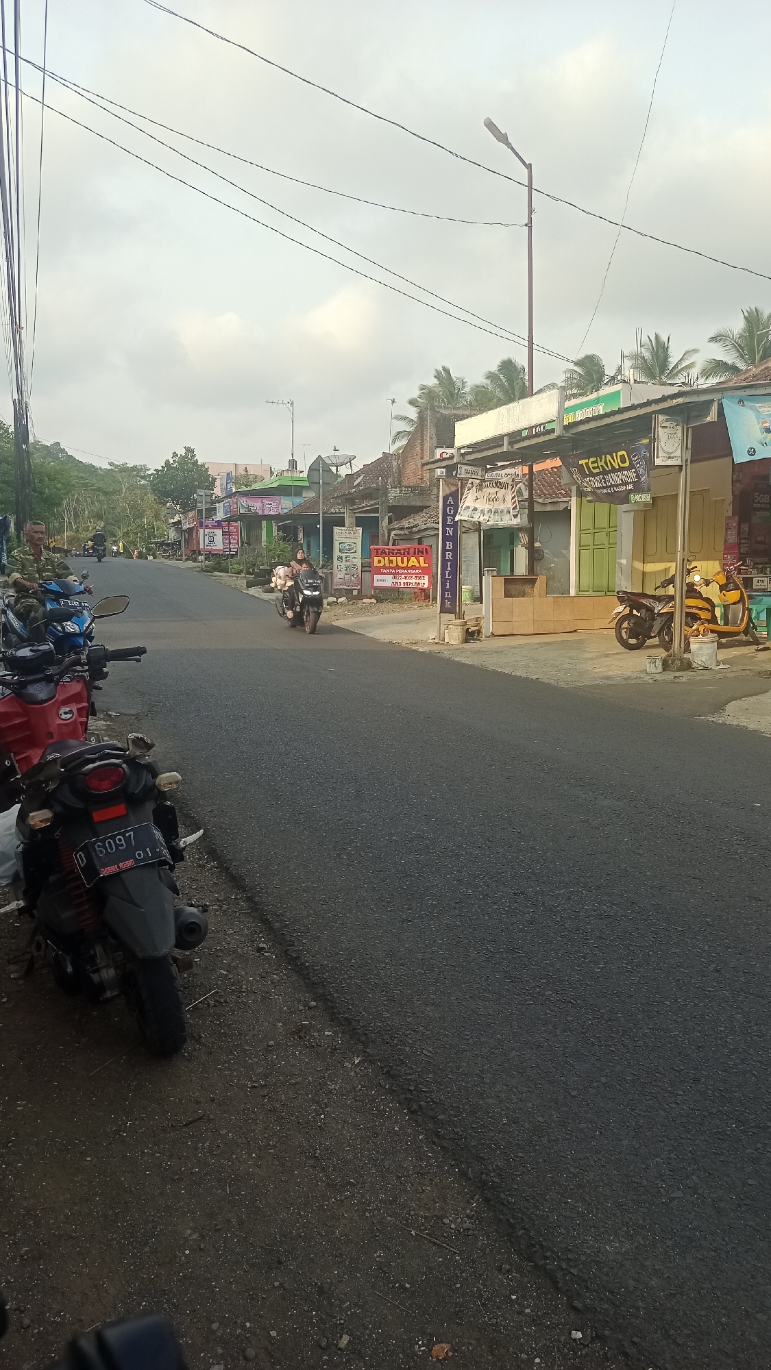 The image depicts a street scene in Karyamandala, Indonesia, likely in the late afternoon or early morning given the lighting. A paved road runs through the frame, with several parked motorcycles on the left side and a few motorbikes in motion on the right. Buildings line the right side of the street, featuring various storefronts with signage, some of which appear to be shops or service providers. One prominent sign reads "TEKNO SERVICE HANDPHONE". A yellow scooter is parked on the sidewalk near a building. The sky is overcast with some clouds visible. The environment appears to be a semi-urban or suburban setting, with a mix of residential and commercial structures.