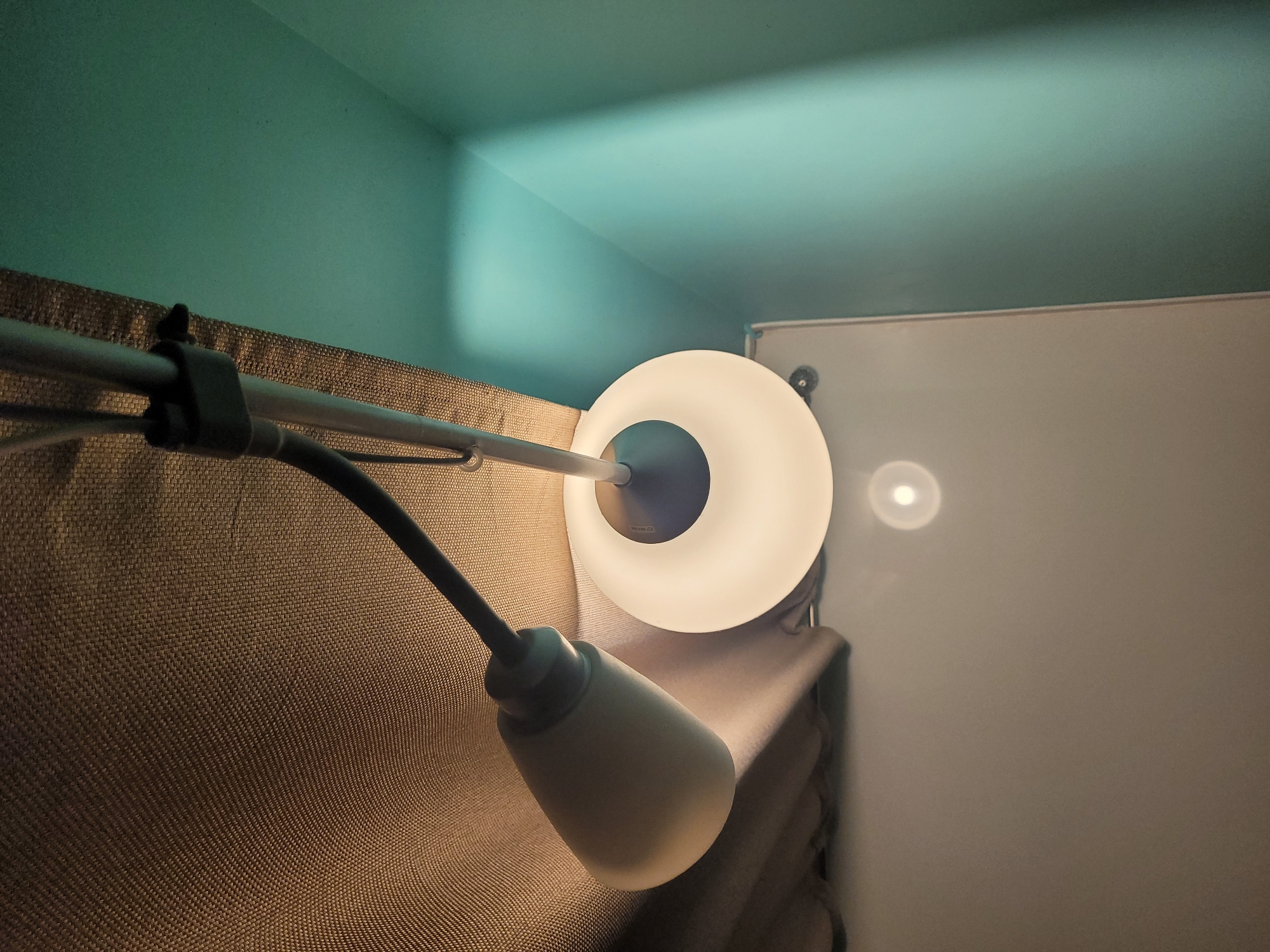 An indoor shot shows a lamp attached to a horizontal rod against a textured wall. The lamp has a round, white, glowing shade and a cone-shaped base. The wall behind the rod is brown and textured. Above the wall is a turquoise-painted area meeting the ceiling. To the right, a white surface is visible, likely a wall or door, reflecting the light from the lamp. There is no indication of this being located in the city of Izhevsk, Russia.