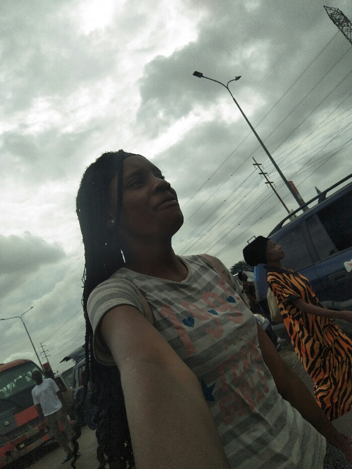 The image captures an outdoor urban scene in Ajah, Nigeria, under a heavily overcast sky. In the foreground, a young Black female with long dark braided hair and a grey and white striped t-shirt featuring pink hearts and blue text is depicted. Her right arm is extended, suggesting she is taking a selfie, and she is looking upwards and slightly to her right.
In the midground, to the right of the primary subject, another young Black female is visible, wearing a black head covering and an orange and black zebra-print dress, also looking upwards. Behind her is a dark blue SUV or van. To the far left, a red and white bus or truck is partially visible, with a man in a white t-shirt standing nearby. Overhead, multiple utility poles with numerous power lines crisscross the frame horizontally, and two streetlights are discernible, one prominently curved in the upper right. A tall metal lattice tower is also visible in the far upper right background.