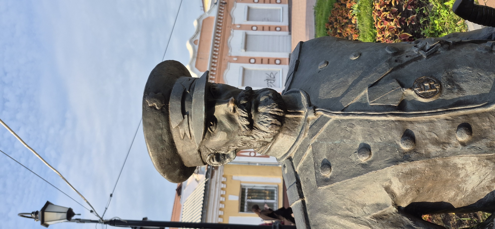 The image is a medium shot, taken outdoors, focusing on a bronze bust of a man in a military uniform. The statue is positioned in front of a building with yellow and pink facades and closed shutters, suggesting an urban setting. The man in the bust wears a peaked cap with a star emblem and a medal on his chest, indicative of a historical military figure. His face is detailed with a mustache and beard, and his expression appears stern. In the background, a lamppost stands to the left, and a person is walking away from the camera on the left side of the frame. Flowerbeds with green grass and red and green foliage are visible to the right of the statue. The sky is partly cloudy and blue. This statue is located in Ussurijsk, Russia, specifically in the central part of the city, known as the Square of the Liberation of Ussuriysk.