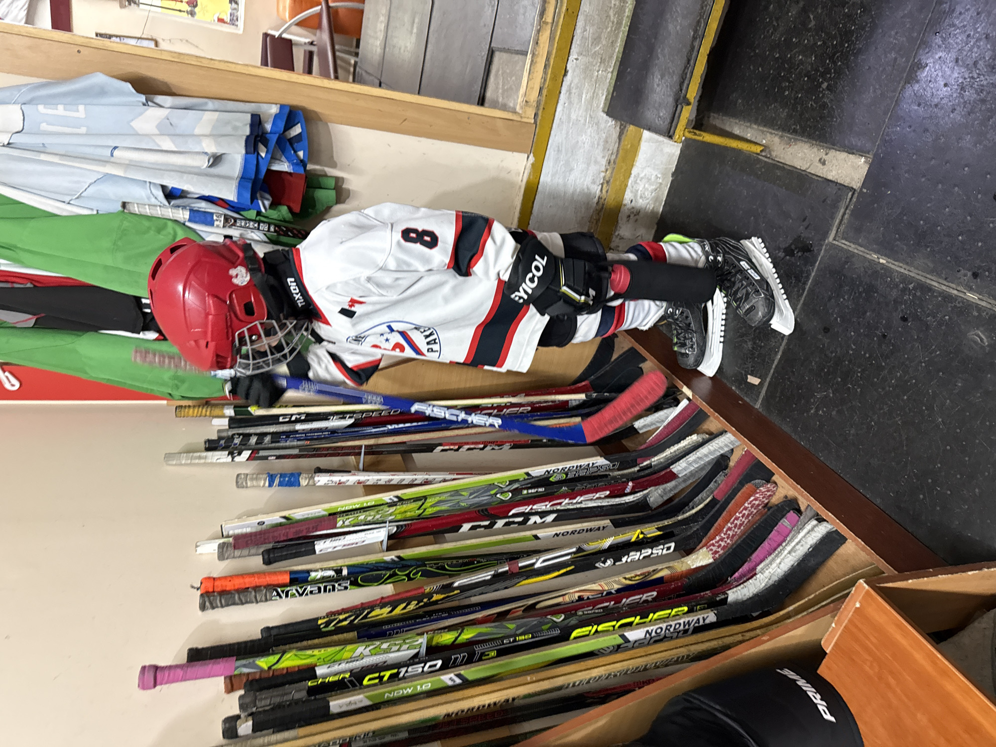 An indoor scene, rotated 90 degrees to the right, captures a young hockey player and an array of hockey sticks in what appears to be a locker room or equipment area in Votkinsk, Russia. A child, likely a boy, is fully dressed in ice hockey gear, including a red helmet with a face cage, a white jersey with black and red stripes and the number '8' on the back, black hockey gloves, and black skates. The child is positioned as if leaning or sitting against a light-colored wall or storage unit, possibly resting or waiting to play.
Above the child, light blue and green folded jerseys are stacked on a shelf. To the right and below the child, a wooden storage rack is filled with numerous hockey sticks, indicating a well-stocked equipment room. The floor is dark, composed of black tiles with some yellow line markings. The setting suggests an area for hockey equipment storage and preparation before or after a game or practice.
Visible text includes: '8' (on the jersey), 'PLAYERS' (on the jersey logo), 'EYICOL' (on gloves), 'TIXON' (on padding), and 'PRIME' (on protective gear). Text visible on various hockey sticks includes 'CM JetSpeed', 'NORDWAY', 'JAPSO', 'FISCHER', 'Arvans', 'CHER', 'CT150', 'NDW 1.0', '36', and 'LE'.
