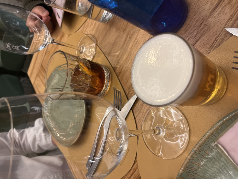 The scene is a close-up view of a table set for four people at a restaurant in Las Palmas de Gran Canaria, Spain, located at the coordinates 28.102 latitude, -15.414 longitude. A beer glass with a white head is in the center of the image. Next to it is a blue glass bottle. To the left, there are two empty wine glasses and a partially visible plate with green patterns on it. The table is made of wood. The place setting is in a casual, yet relaxed dining environment.